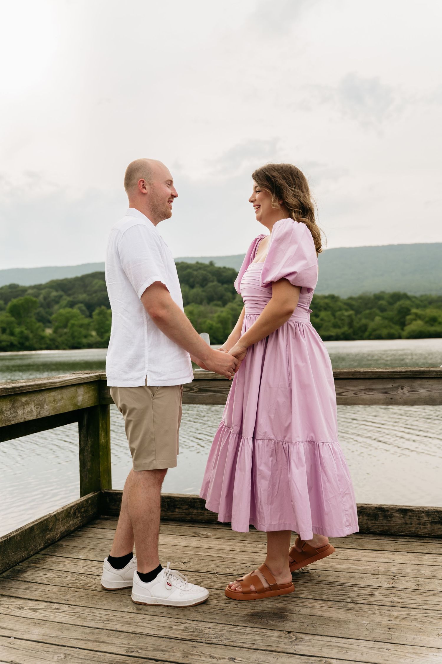 A couple stands holding hands on a wooden deck overlooking rolling green hills on a cloudy day.