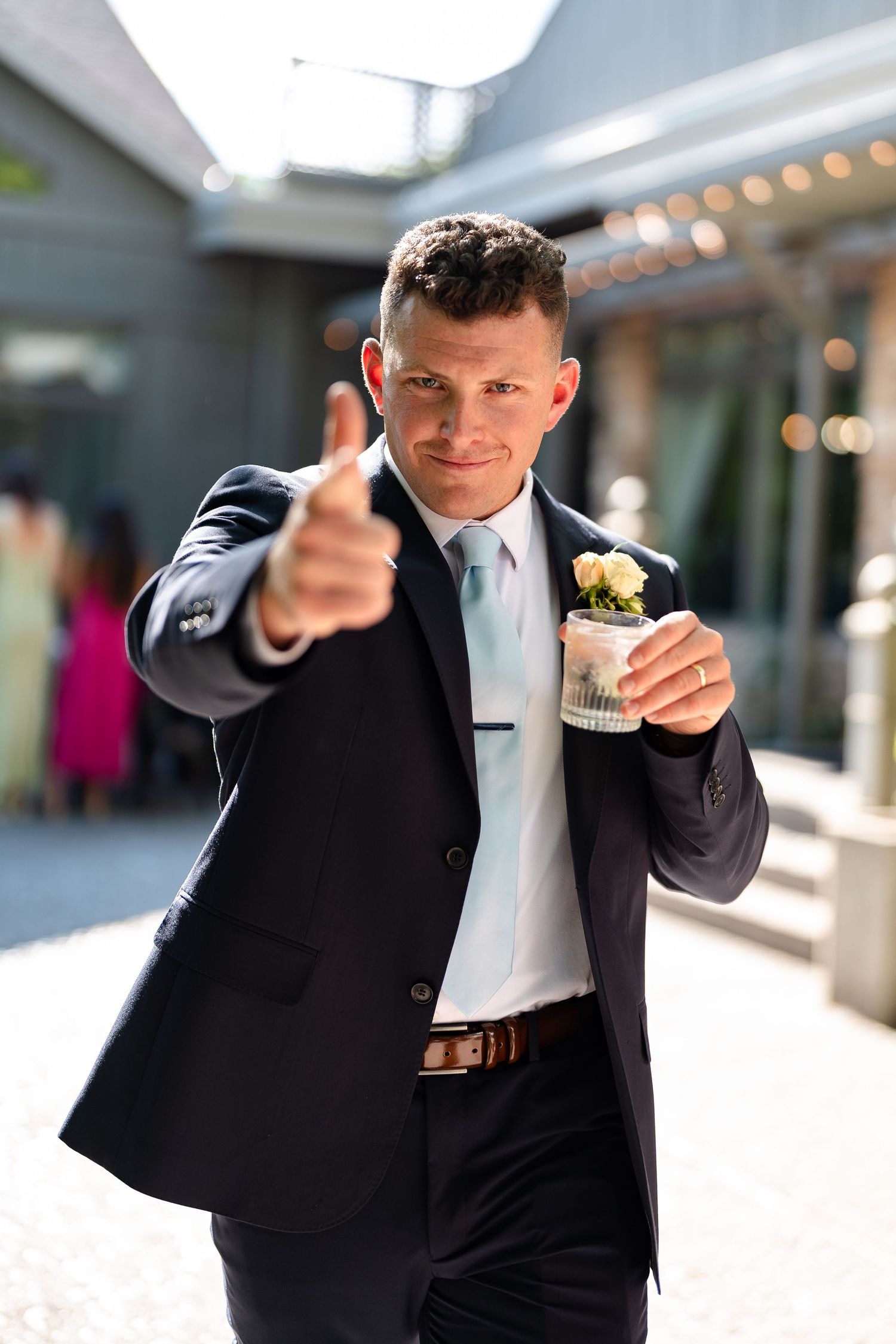 Groom in navy suit giving thumbs up while holding drink with boutonniere at outdoor wedding venue.