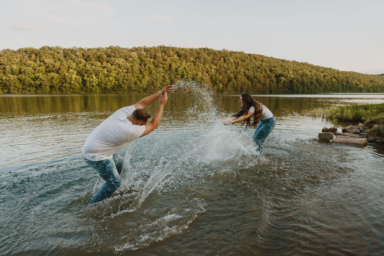 Two people playfully splash water at each other in a scenic lake.