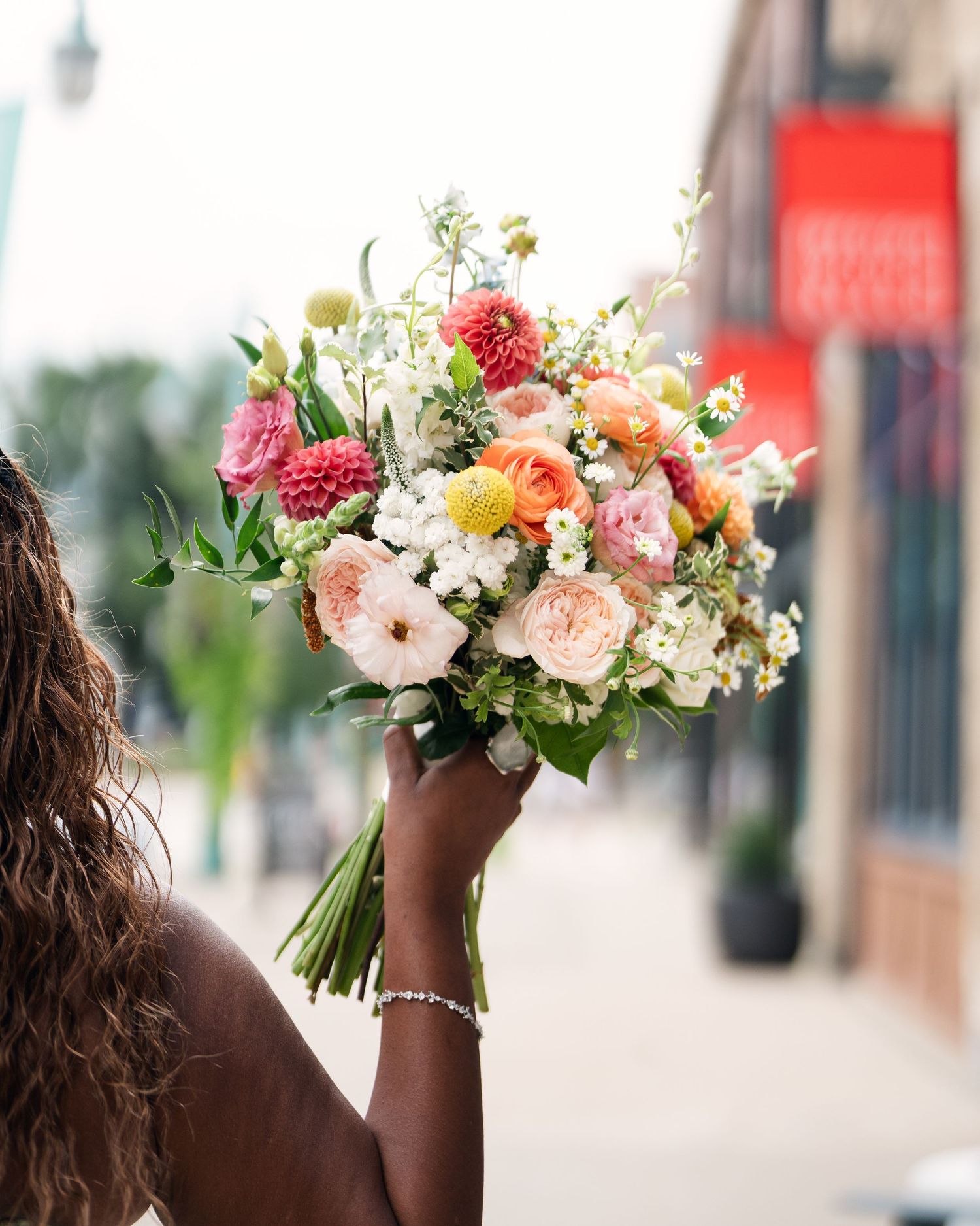 A bride holds a vibrant bouquet of coral, pink, and white flowers aloft on a city street.