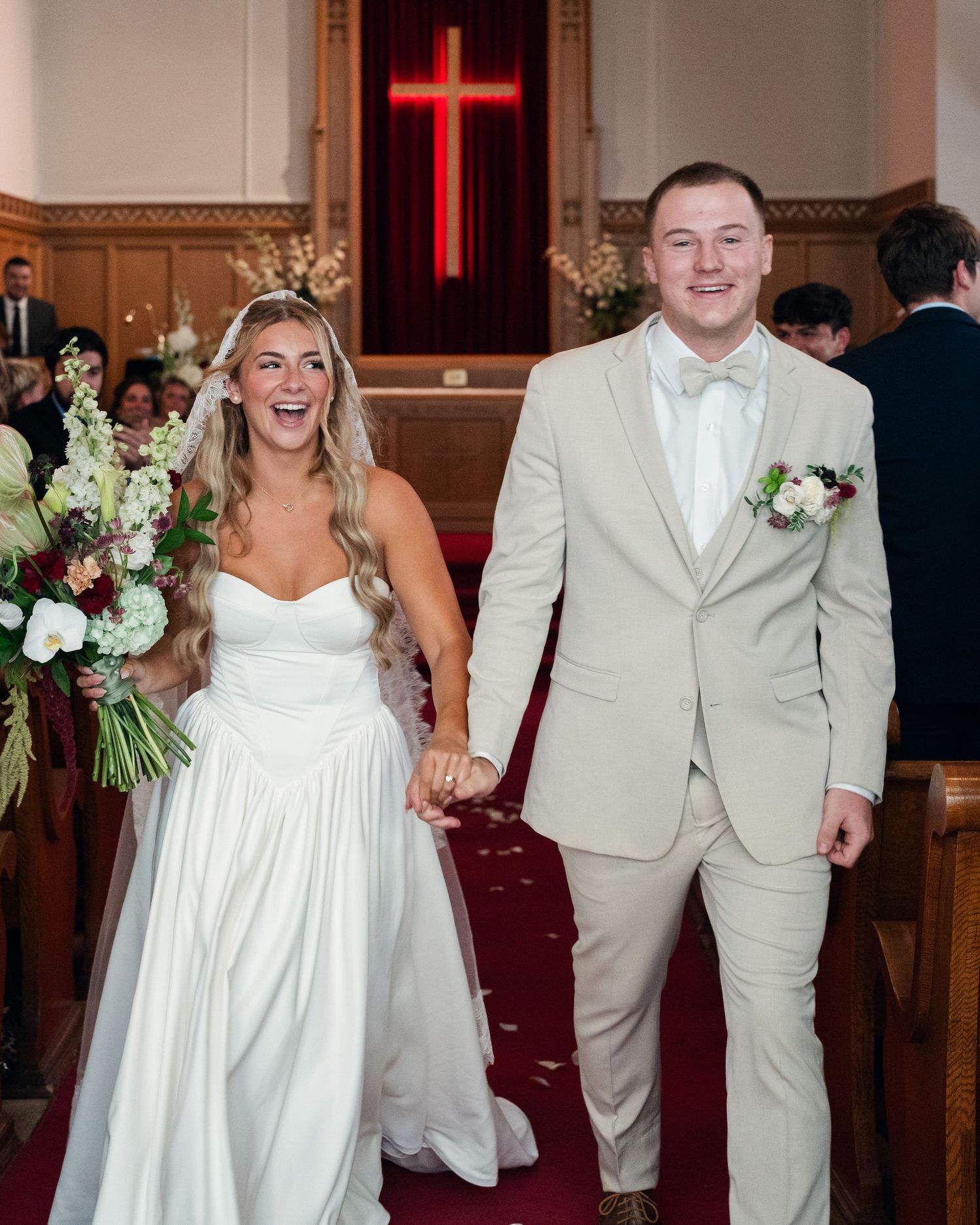Joyful bride in white gown and groom in tan suit walk down church aisle after wedding ceremony.