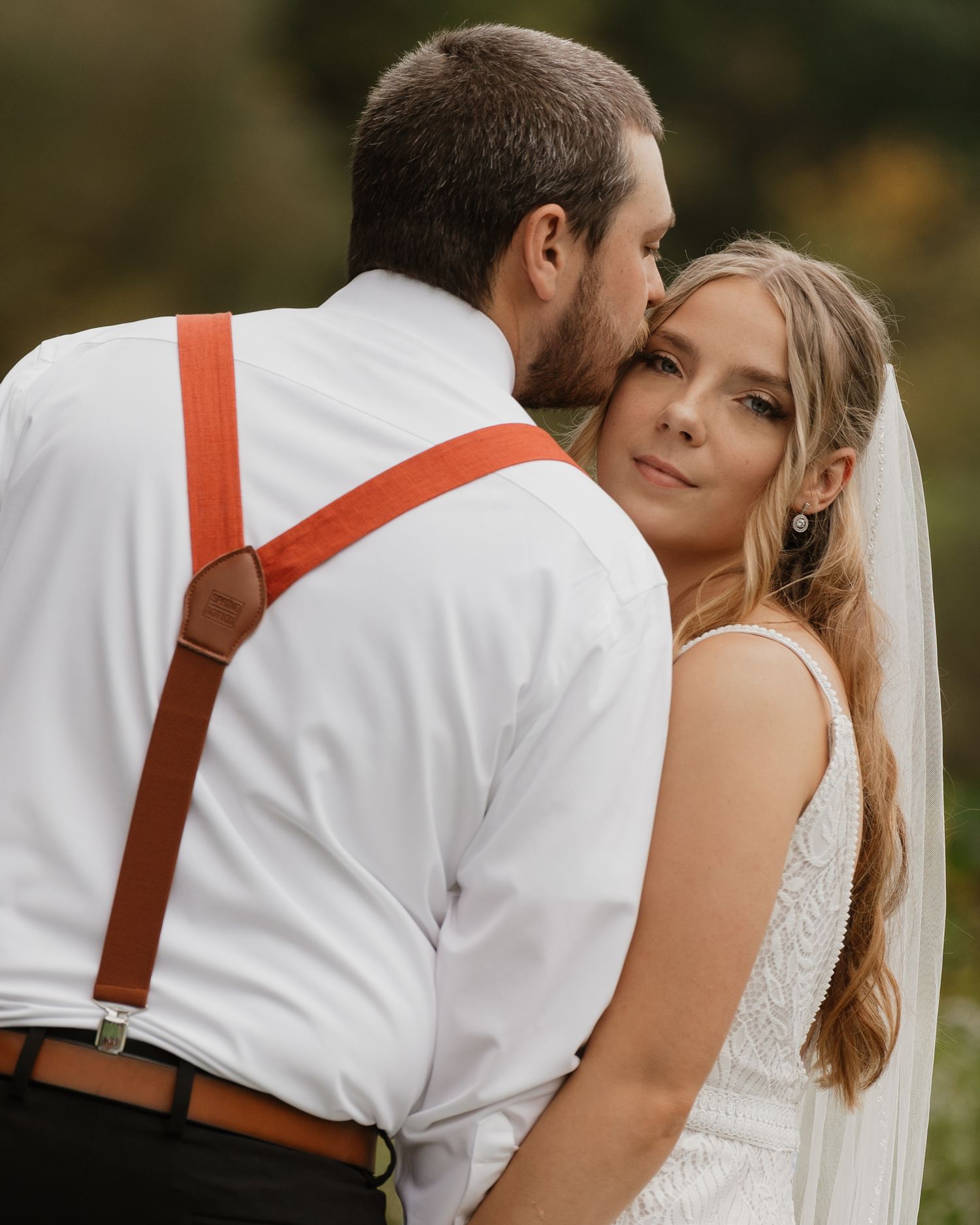 Wedding couple in formal attire embracing outdoors with brown leather suspenders detail.