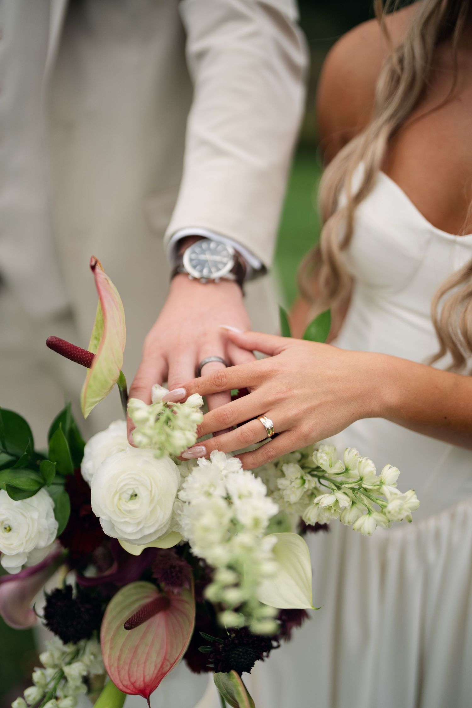 Bride and groom displaying wedding rings over a bouquet of white ranunculus and burgundy anthuriums.
