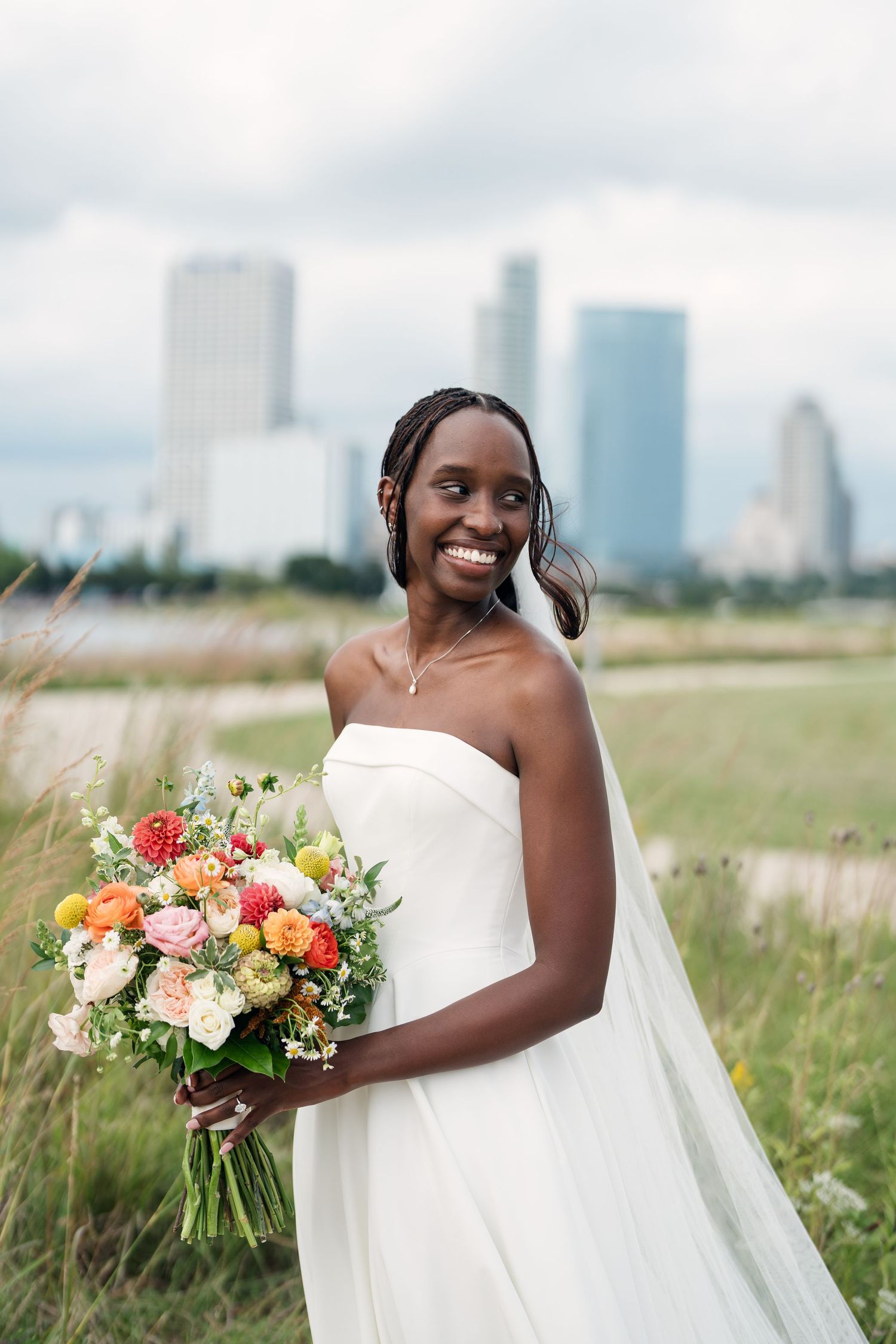 Smiling Black bride in strapless white gown holds colorful bouquet in field with city skyline backdrop.