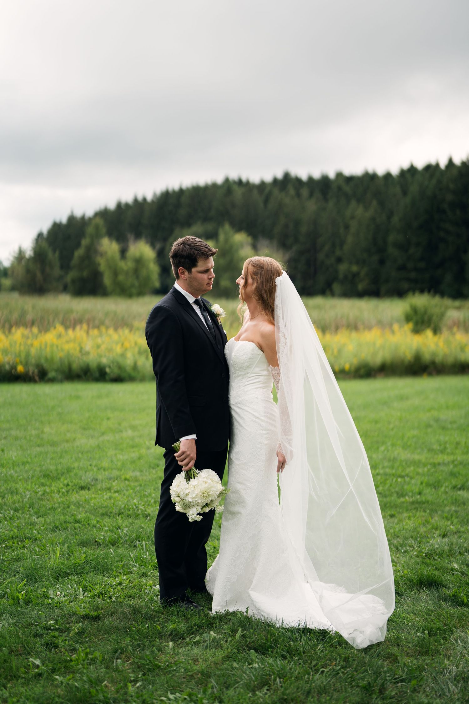 Bride and groom standing face to face on green grass with forest backdrop on their wedding day.