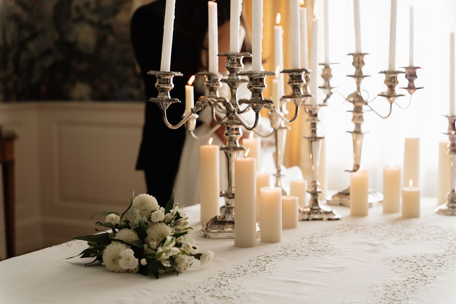 Elegant wedding table with silver candelabras, white pillar candles, and a floral arrangement on a lace tablecloth.
