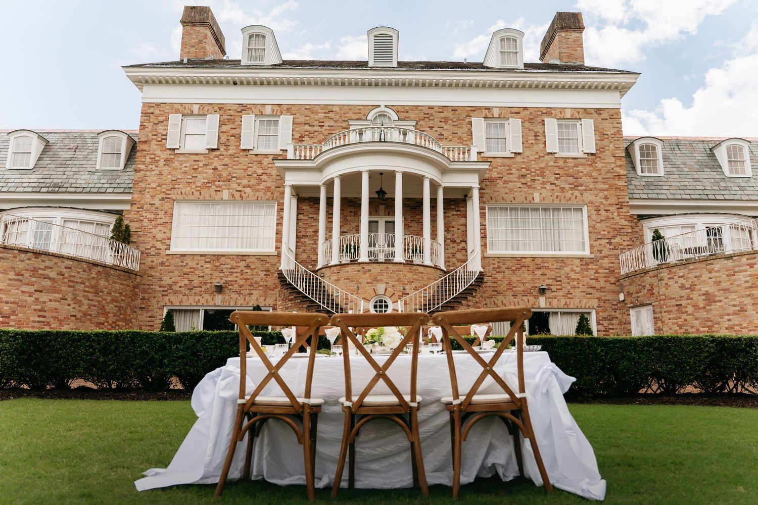 A white wedding tent with cross-back chairs set up on a manicured lawn next to a historic brick mansion.