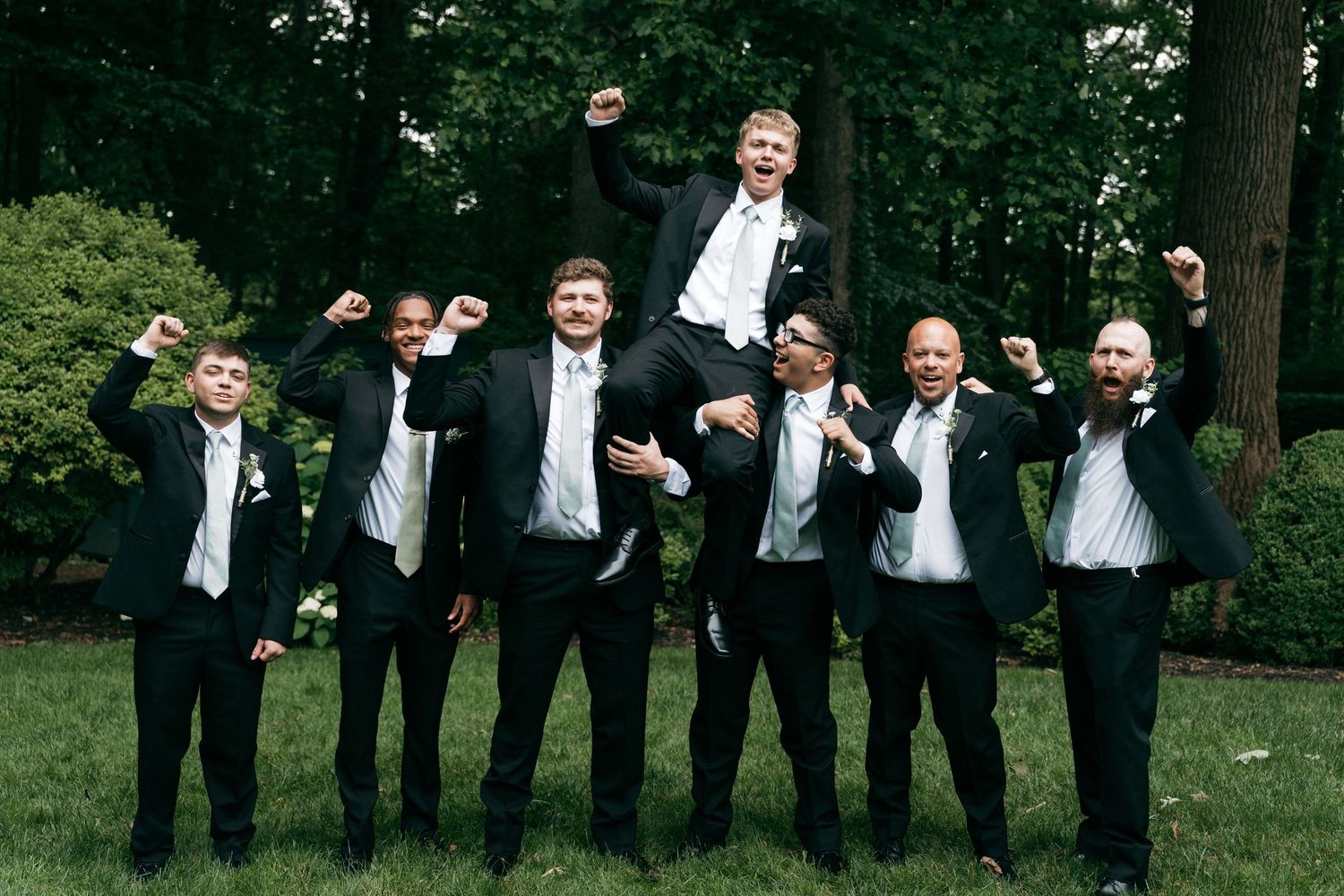 Groom lifted by groomsmen in black tuxedos, celebrating outdoors with raised fists and smiles on a green lawn.