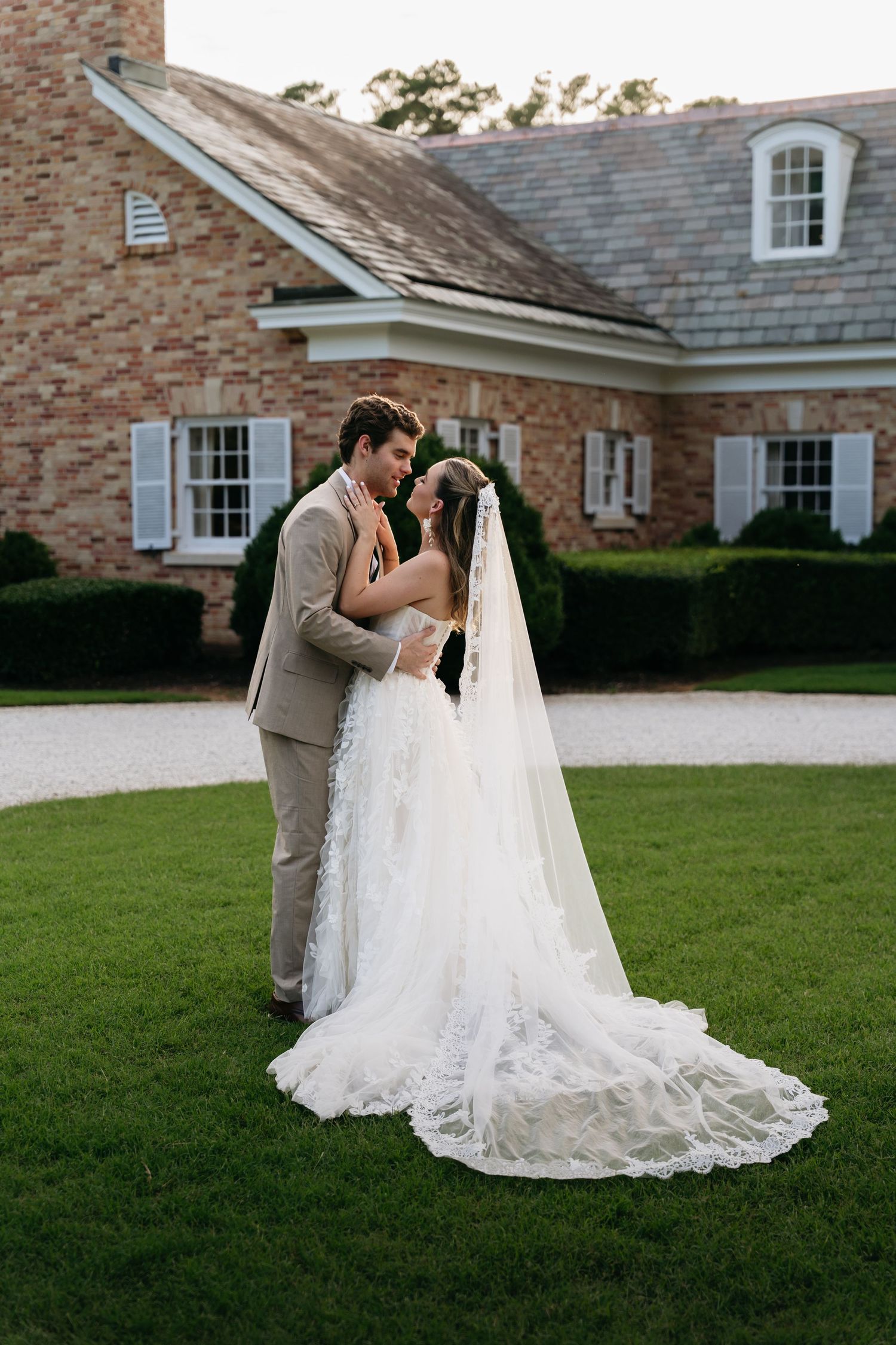 A couple in wedding attire share romantic moments on a lawn outside a brick building with white trim.