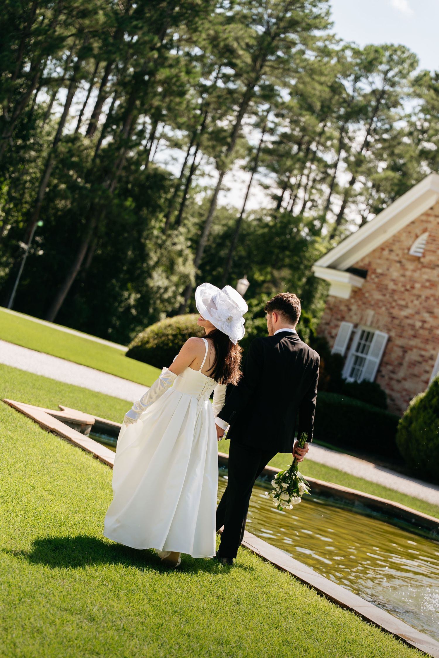 A couple in wedding attire walks together on a green lawn beside a pond on their special day.