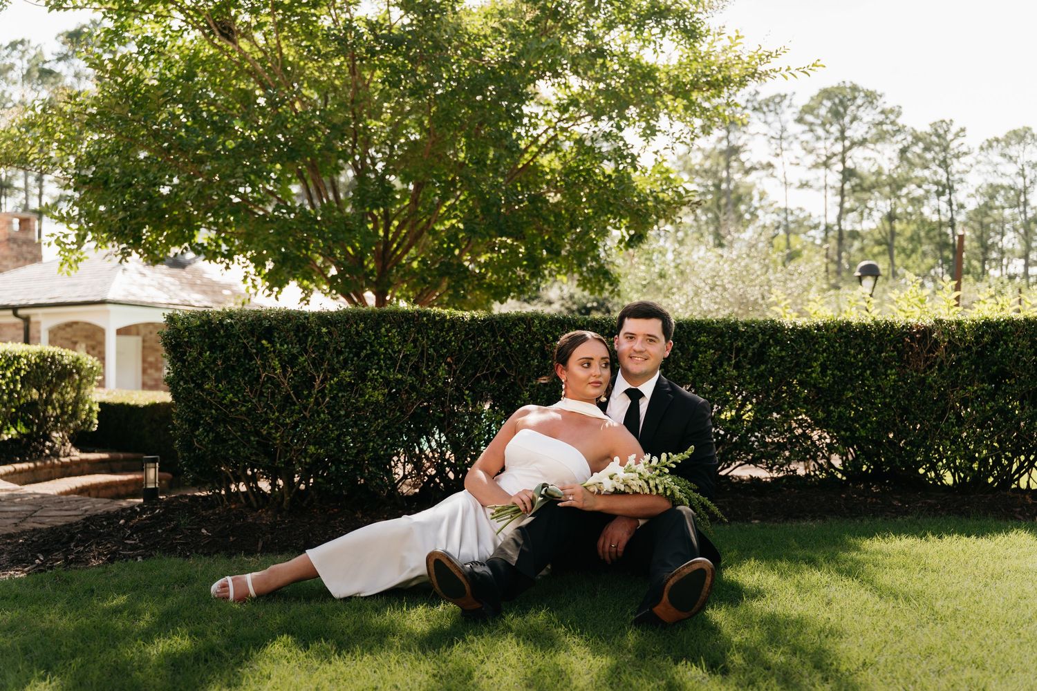 A couple playfully dips and poses together on a sunny lawn surrounded by green trees and hedges.