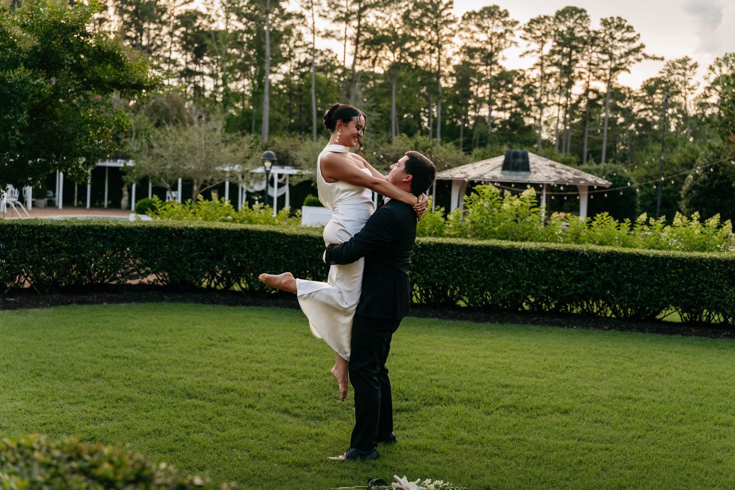 A couple embraces in a romantic lift pose on a manicured lawn with pine trees and gazebos in the background.
