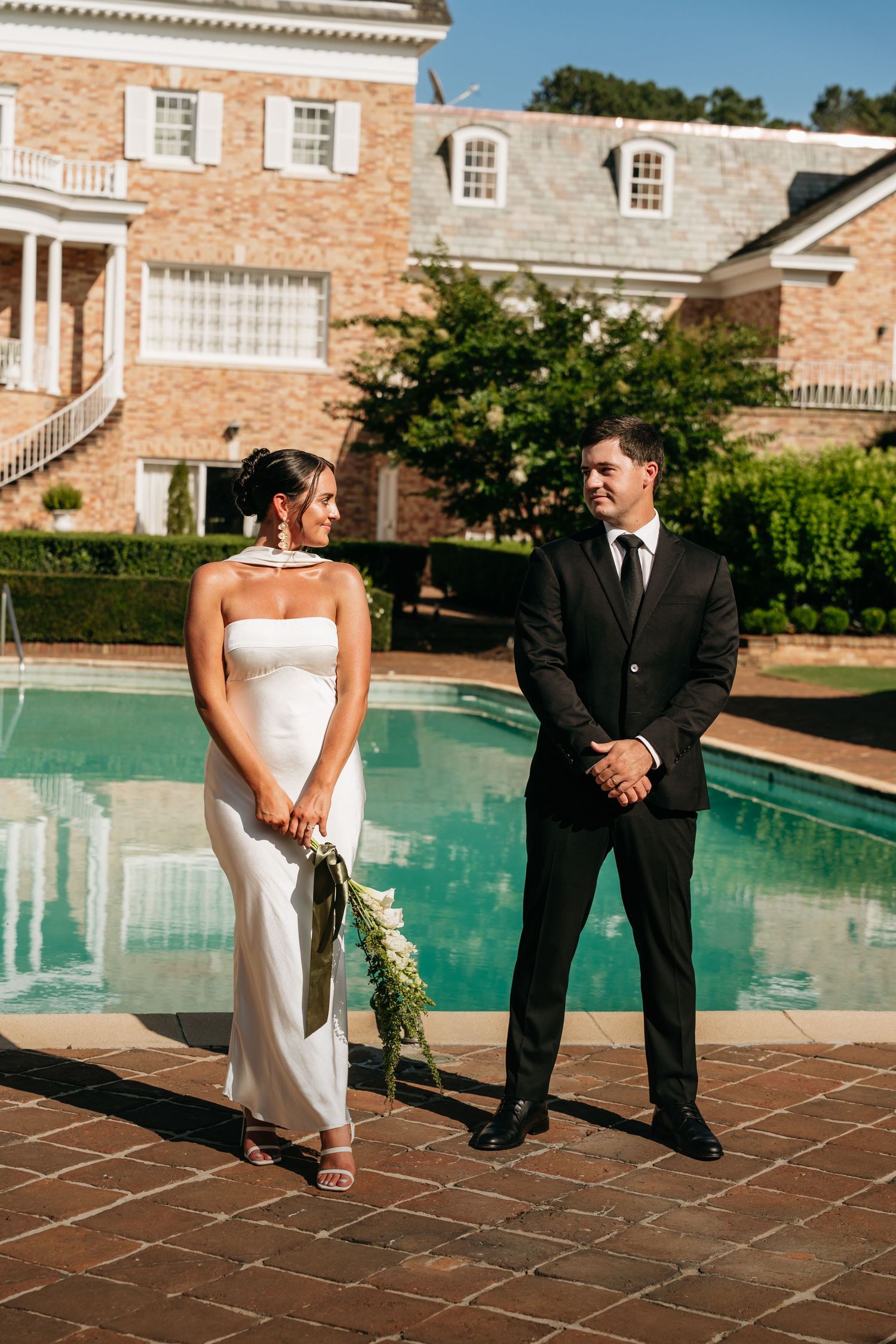 A couple in wedding attire poses playfully by an elegant swimming pool at a brick mansion venue.