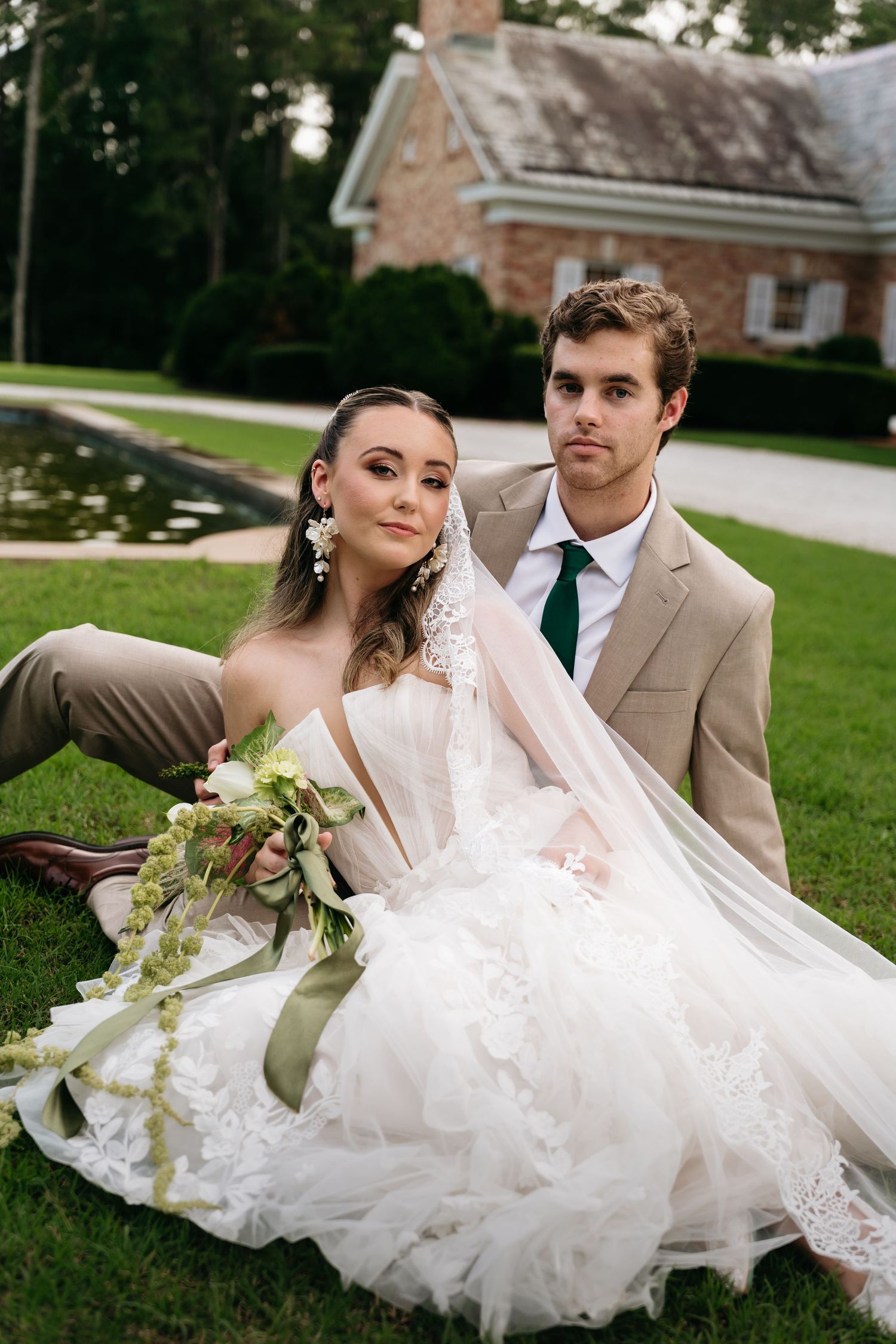 A romantic wedding couple lounges on grass in a series of elegant outdoor portraits with flowing white dress and tan suit.