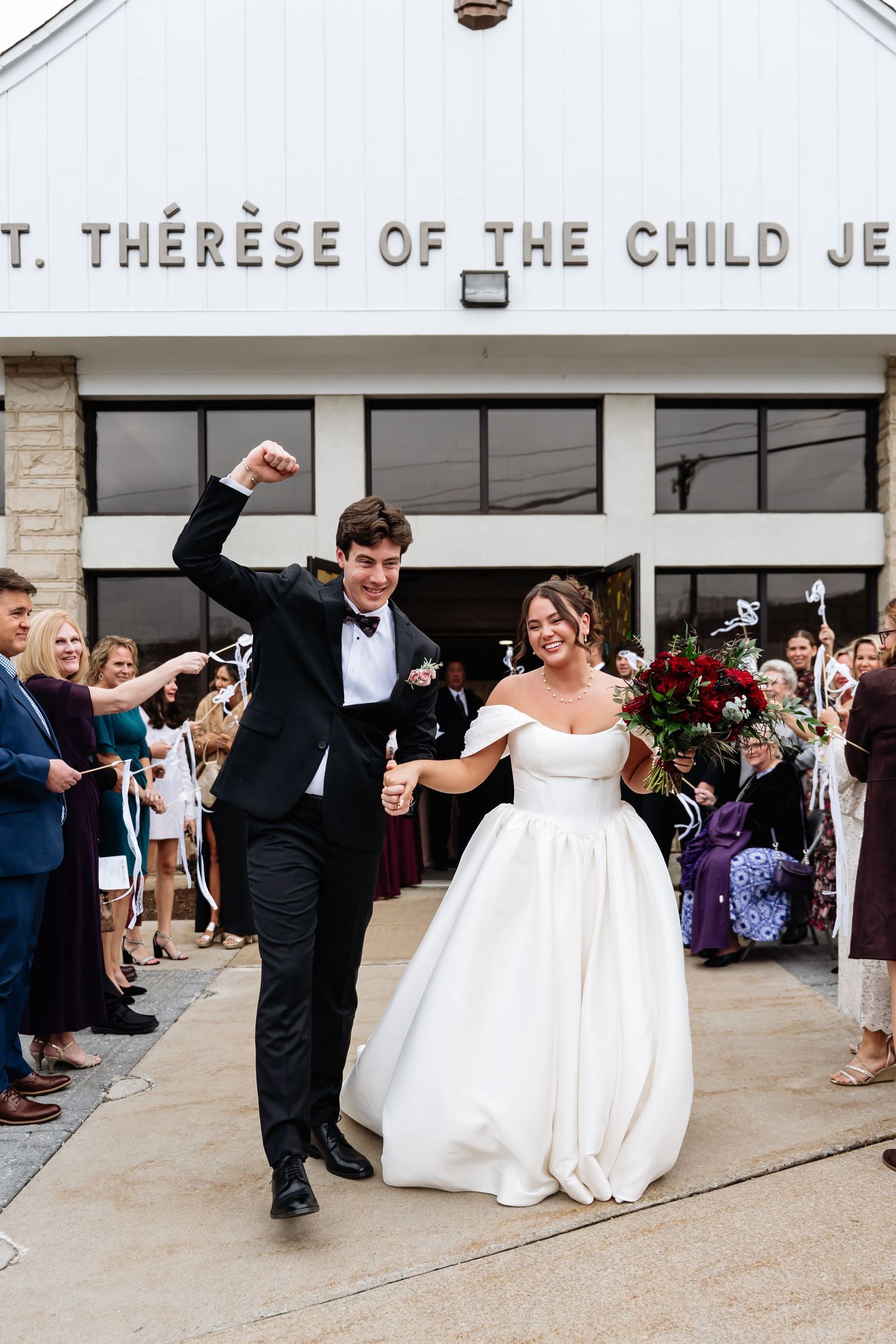 Joyful newlyweds exit St. Therese of the Child Jesus church, groom raising fist in celebration, bride in white gown holding bouquet.