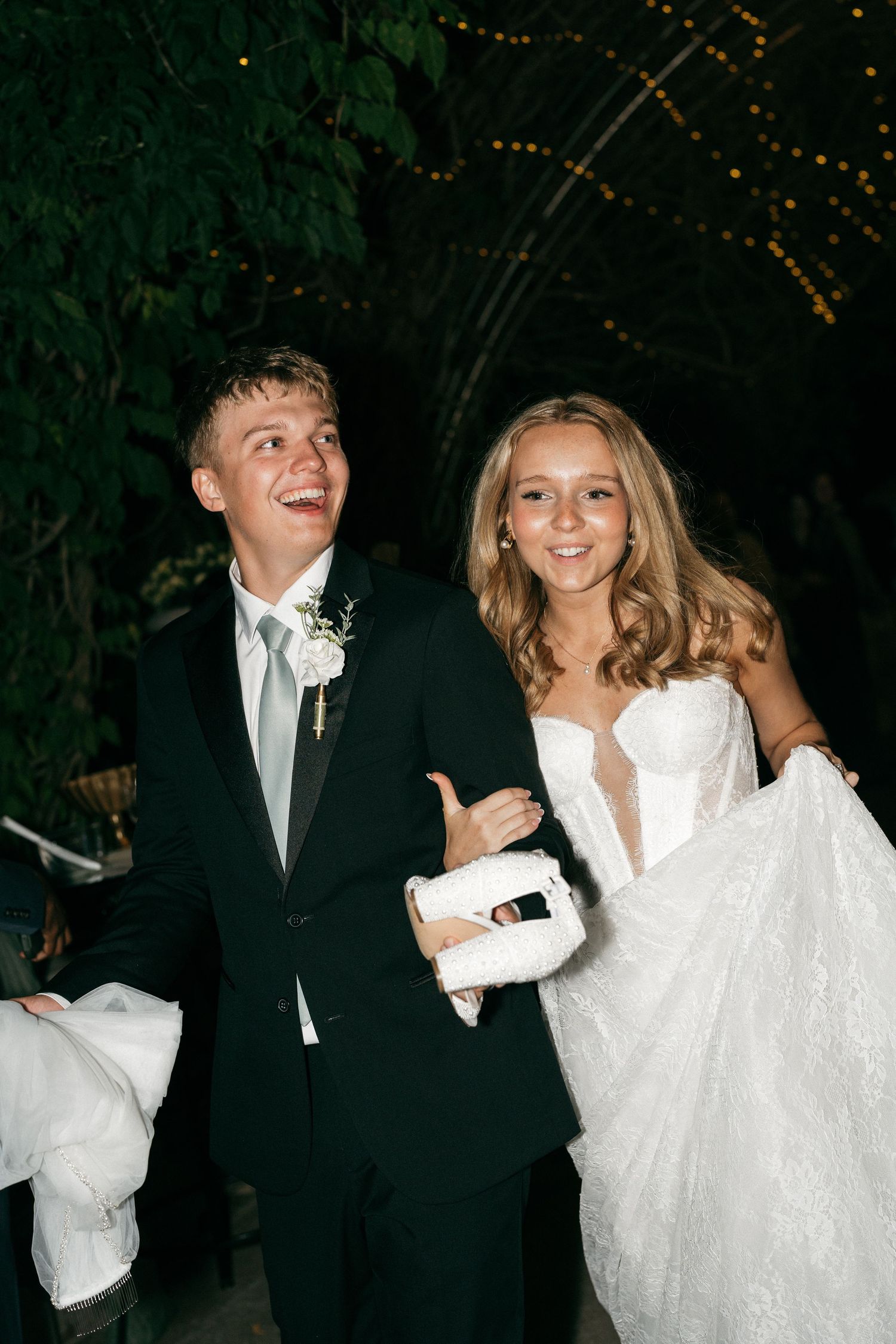 Young couple in formal wedding attire smiling outdoors at night, groom in black suit and bride in white lace gown.