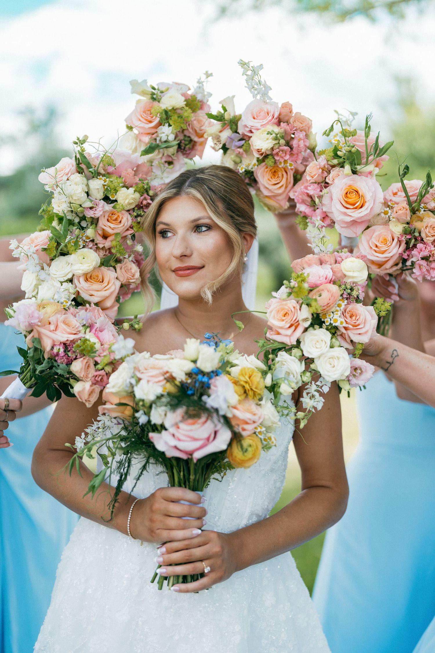 Bride in white gown holding floral bouquet framed by bridesmaids bouquets of pink and peach roses outdoors.