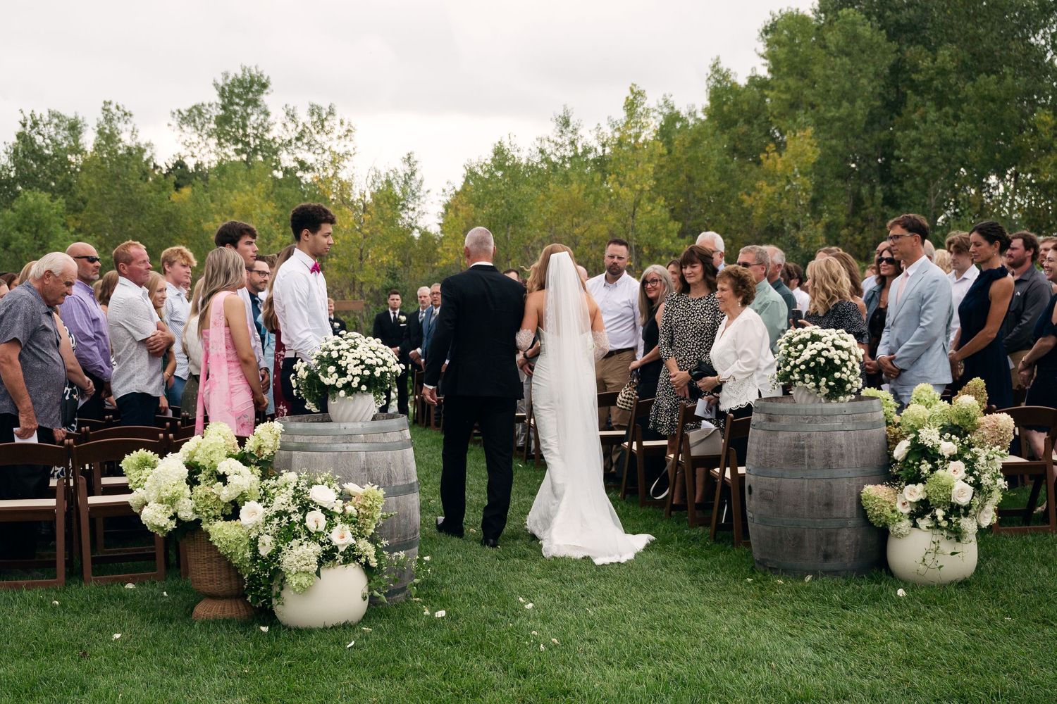 Outdoor wedding ceremony on green lawn with barrel floral arrangements and guests lining the aisle.