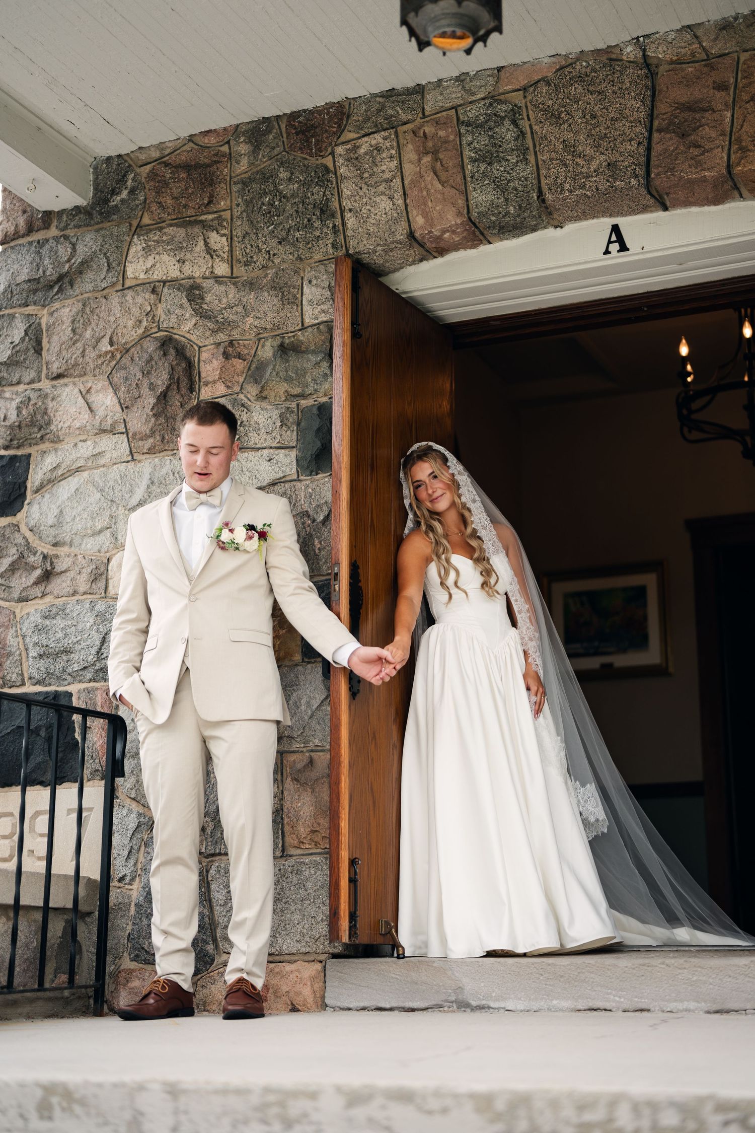 Bride and groom hold hands at stone building entrance, groom in beige suit, bride in white gown with veil.