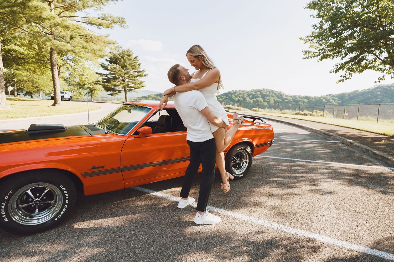 Playful couple poses with orange vintage car outdoors near water.