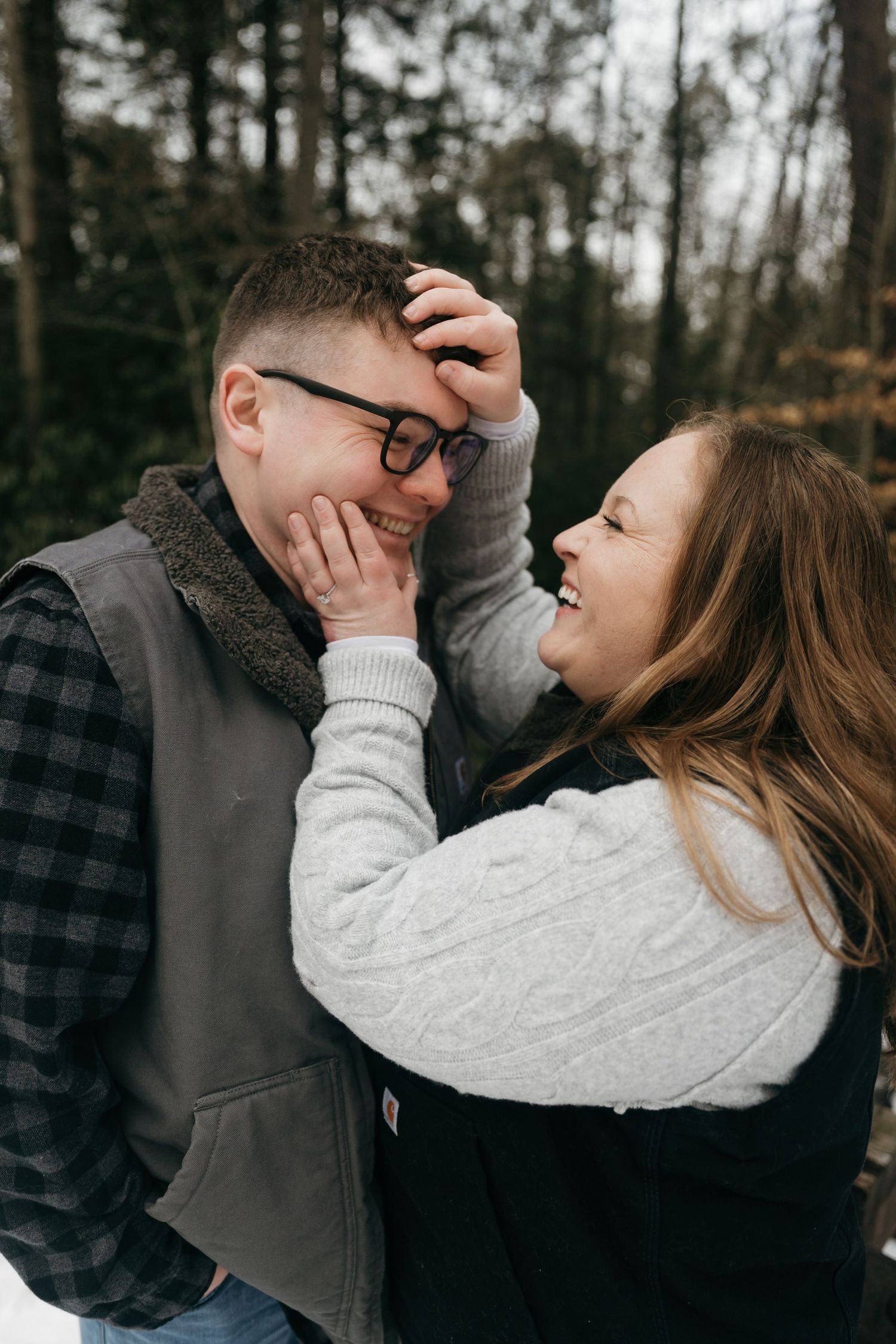 A playful couple laughing and touching faces in a winter forest setting.