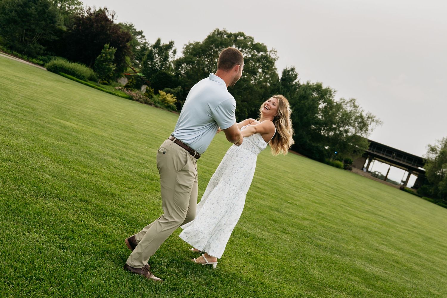 Couple dancing on lush green lawn, man in blue shirt twirling woman in white dress outdoors.