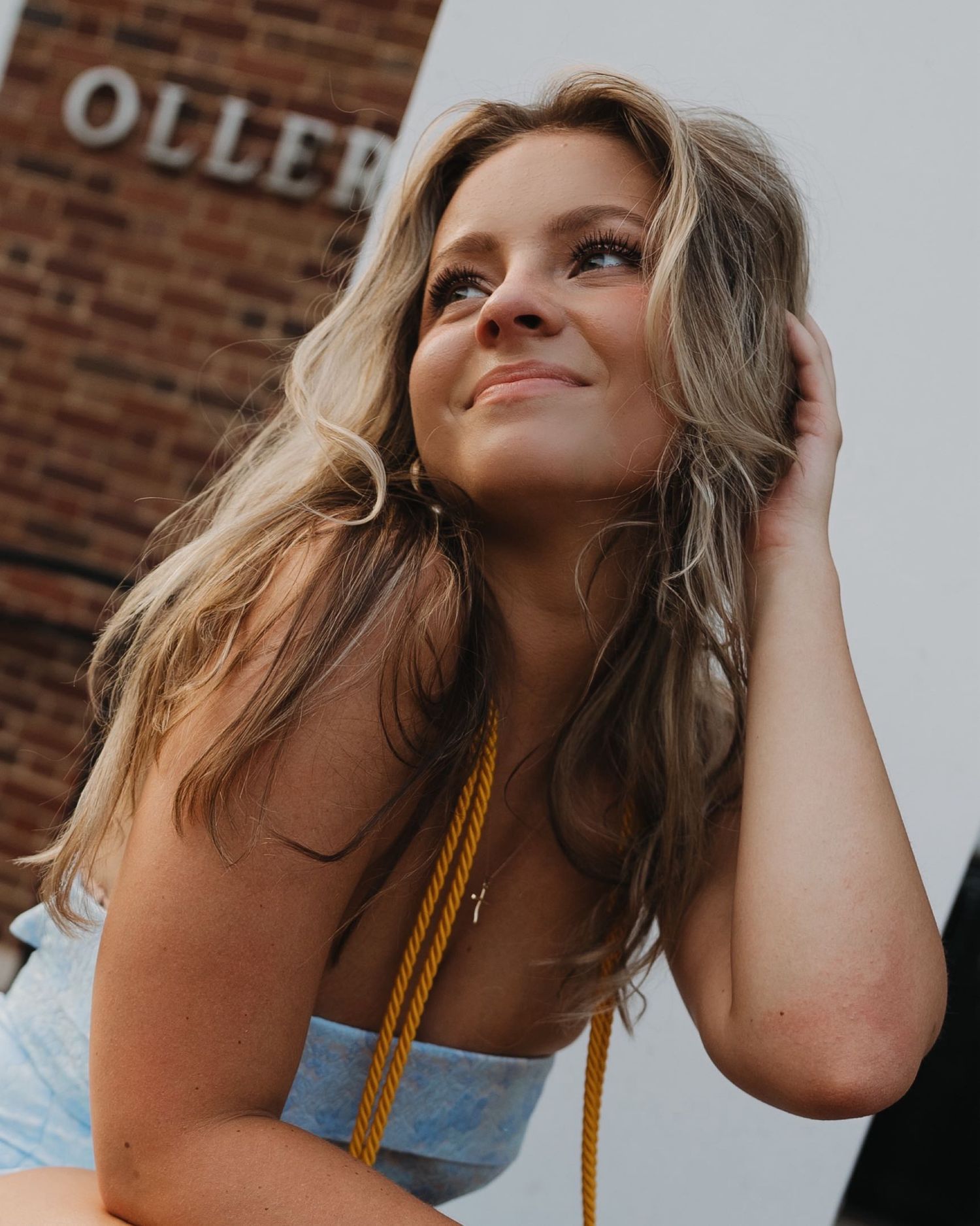Portrait of a woman with long blonde hair smiling against a brick wall with vintage signage.