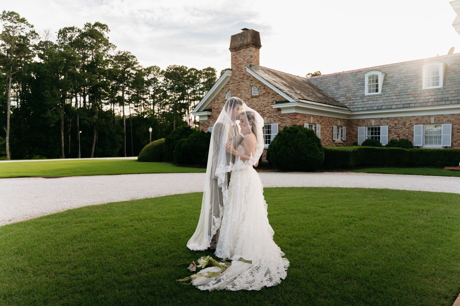 Bride and groom share a romantic kiss on lush green lawn outside elegant brick wedding venue at golden hour.