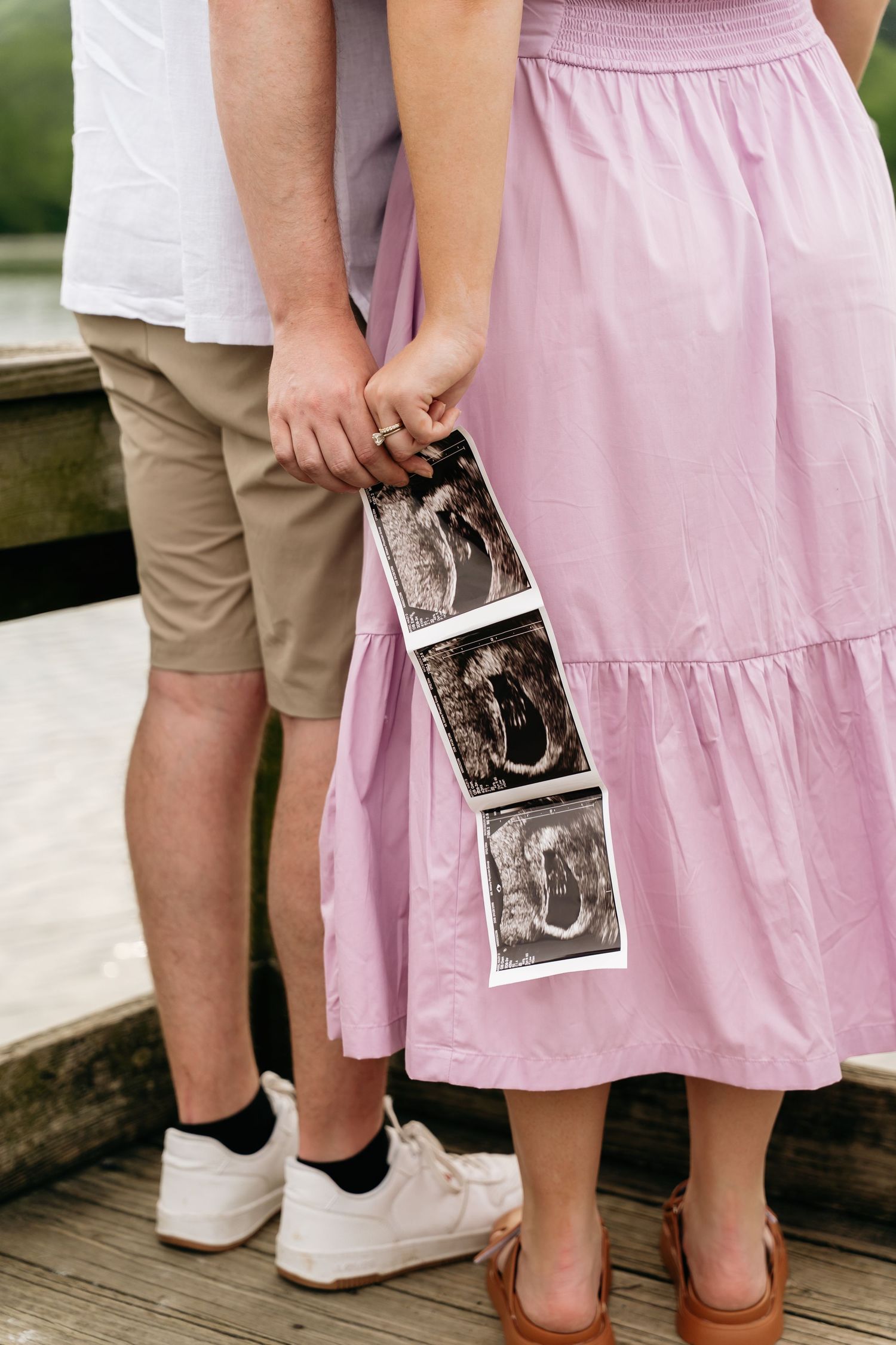 Couple in casual clothing holding ultrasound sonogram photos on a wooden deck in pink dress and khaki shorts.