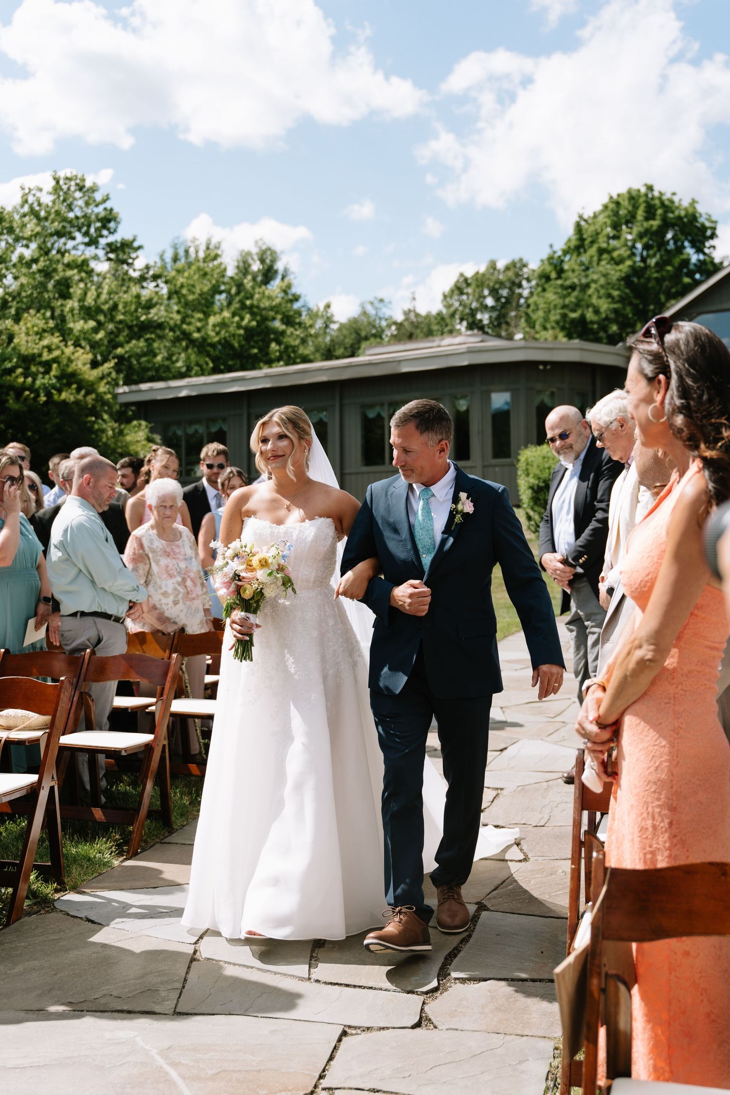 Bride walks down the aisle with her father at an outdoor wedding ceremony on a sunny day.