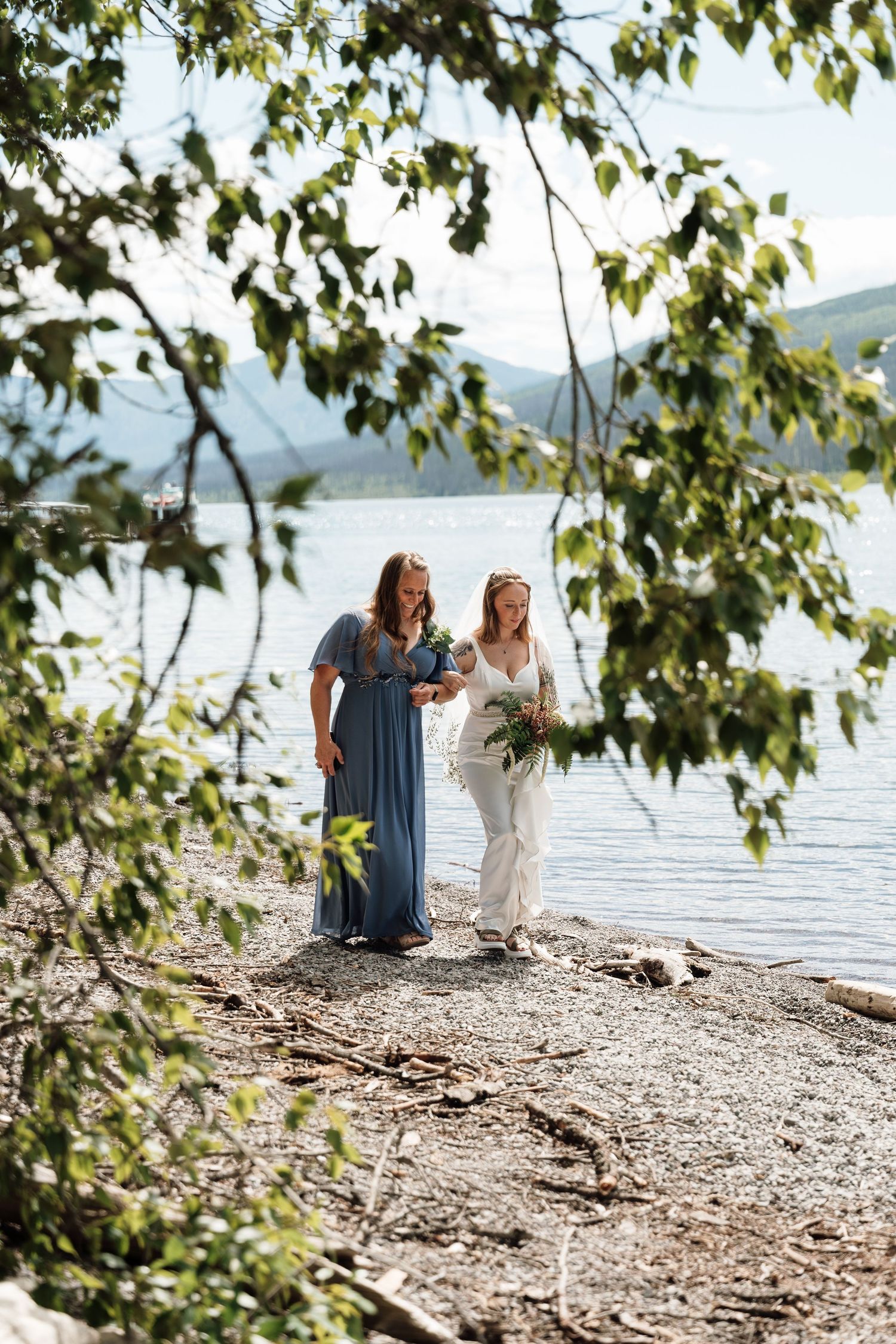 Two women in elegant attire walk along a rocky lakeside shore surrounded by lush green trees.