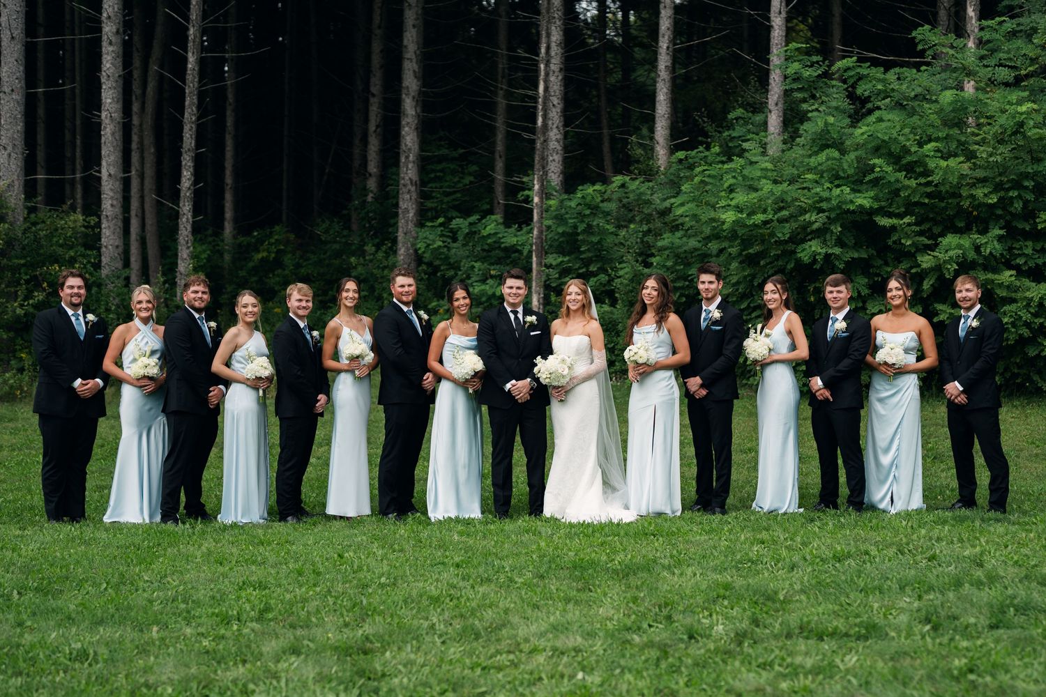 Wedding party poses outdoors on green grass with tall pine trees, bridesmaids in mint gowns and groomsmen in dark suits.