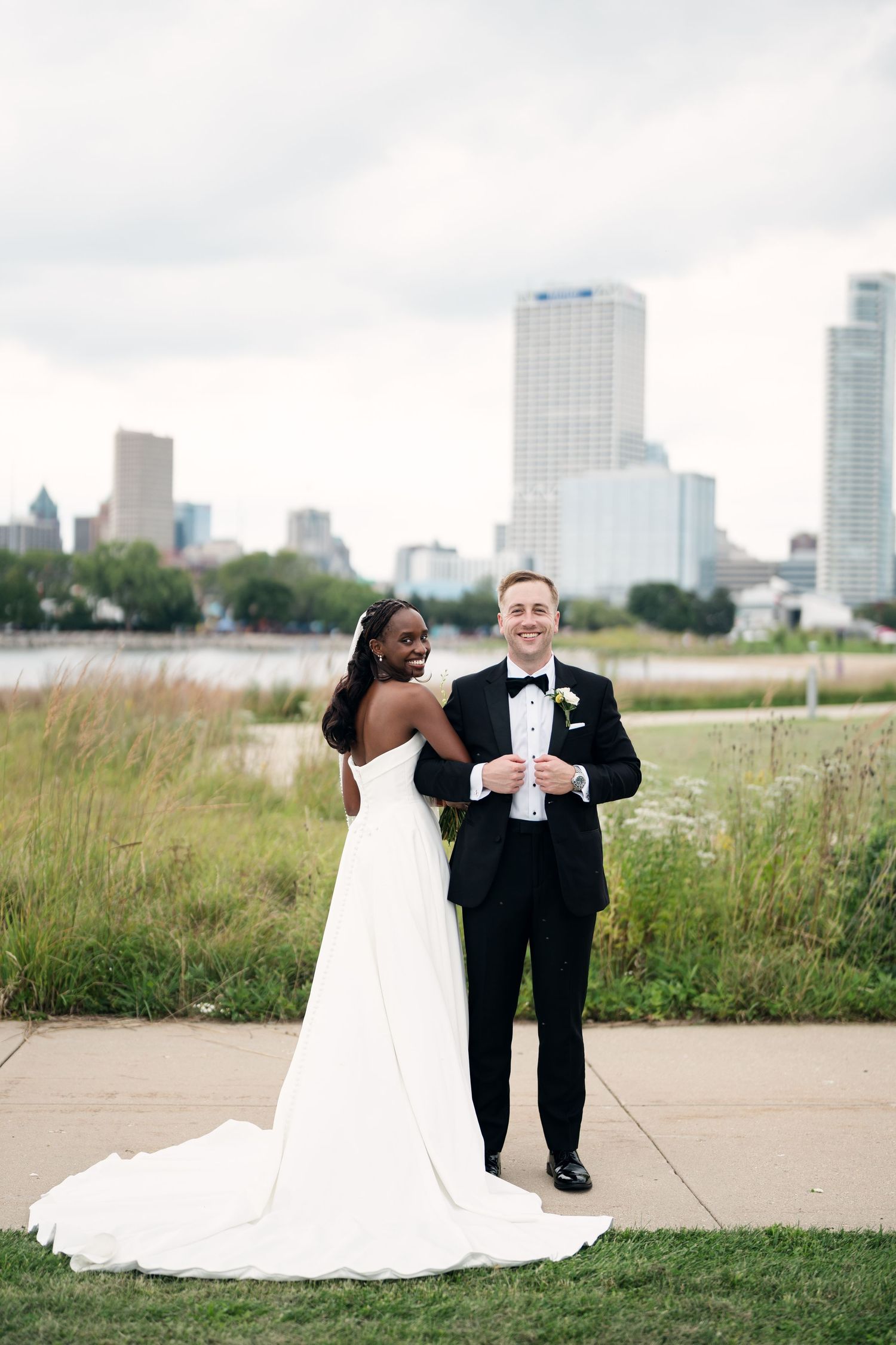 Interracial couple poses in wedding attire with city skyline backdrop, bride in white gown, groom in black tuxedo.
