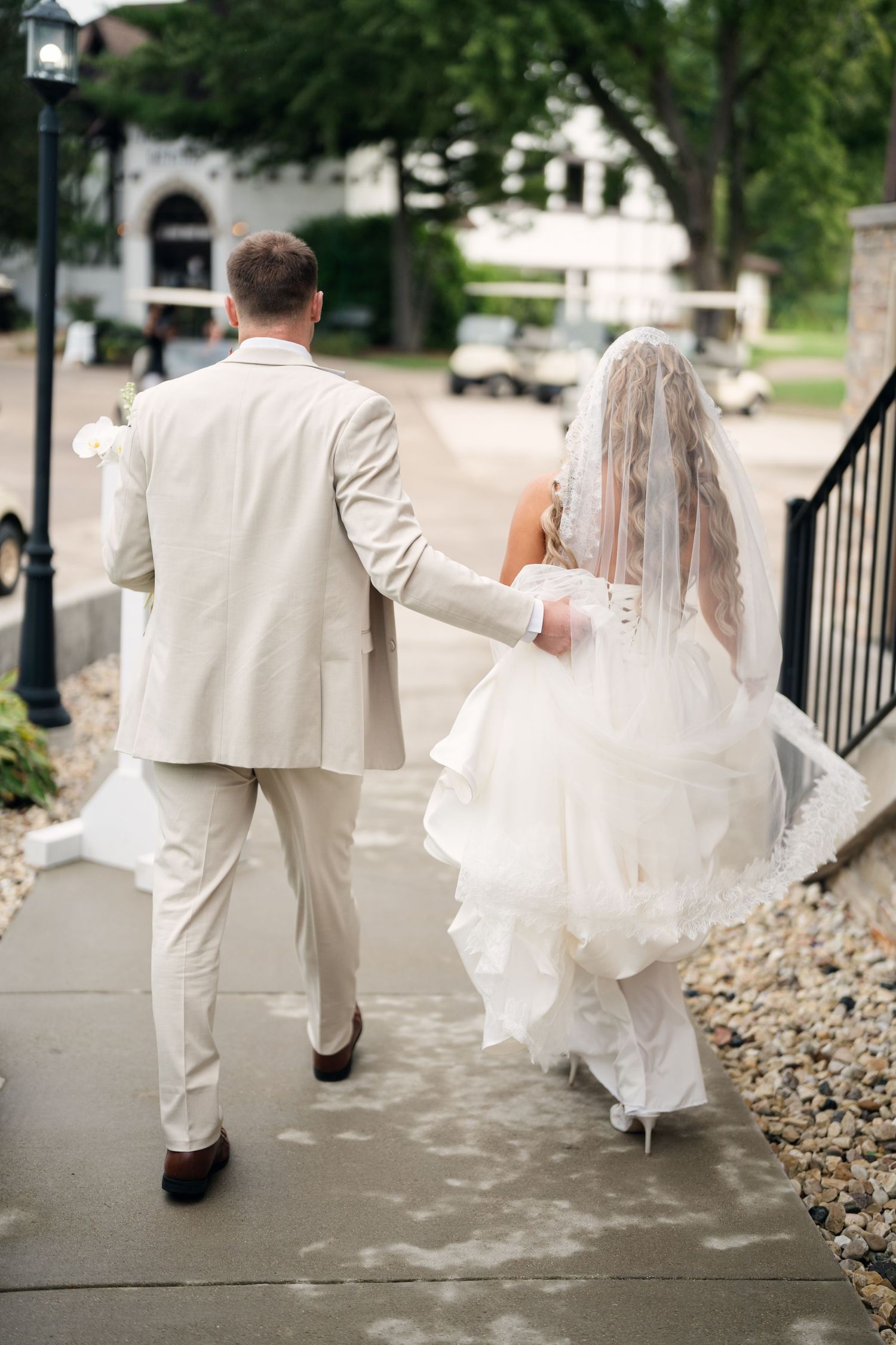 Bride and groom walk hand in hand along a path, bride in layered white gown with veil trailing behind.