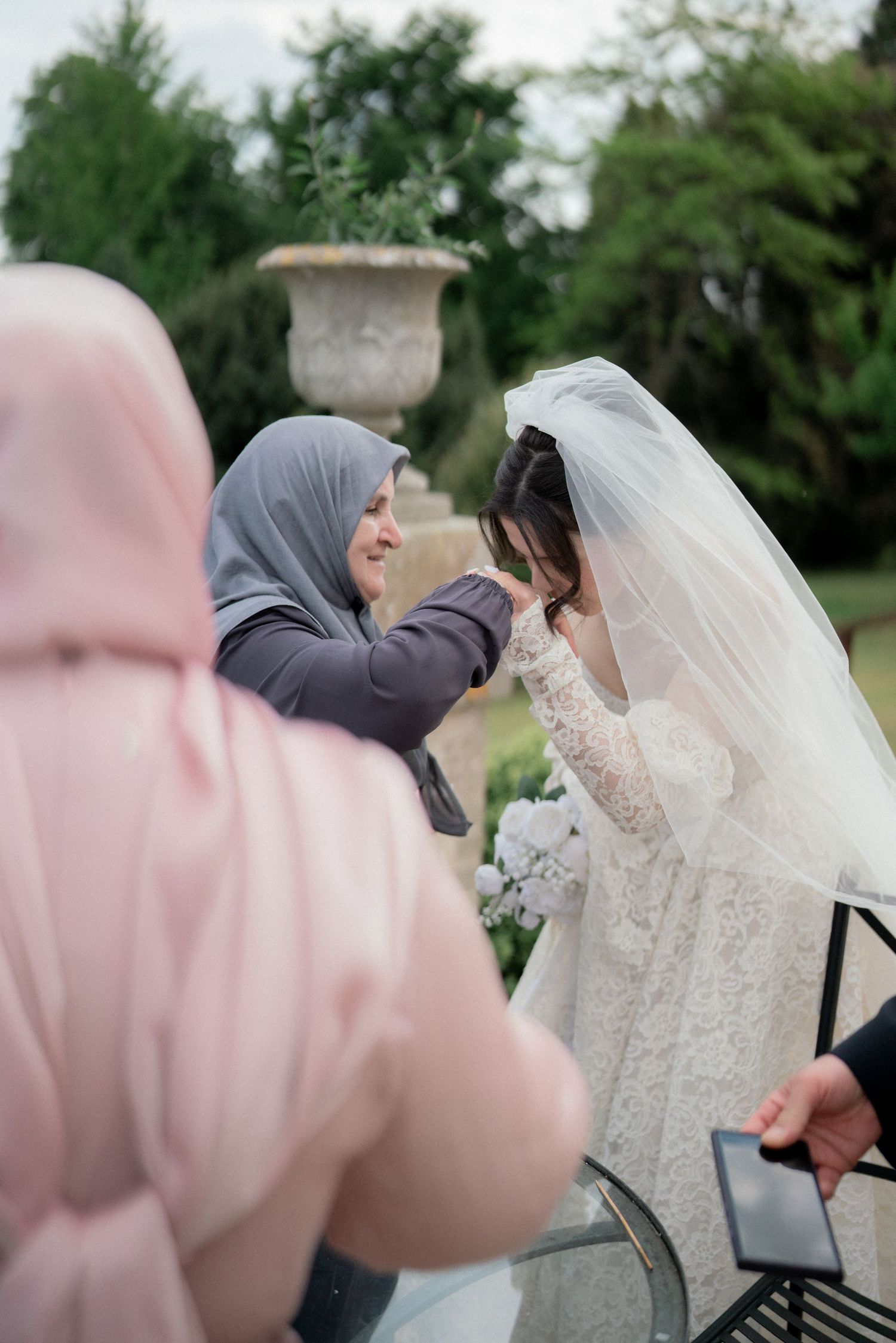 Wedding party members in pink dresses gather around bride in white lace gown outdoors.