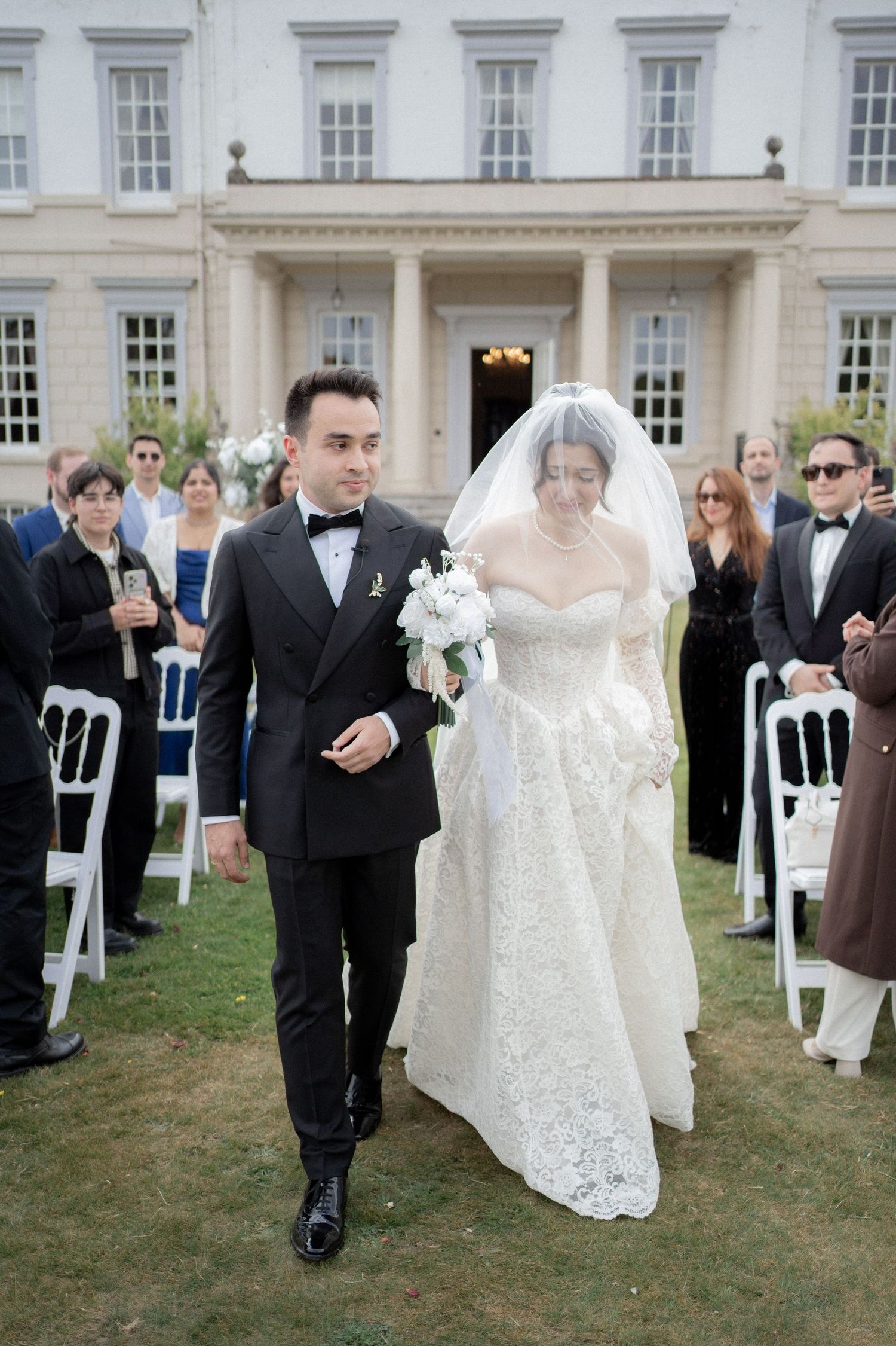 Wedding ceremony procession outside a grand white colonial-style building.