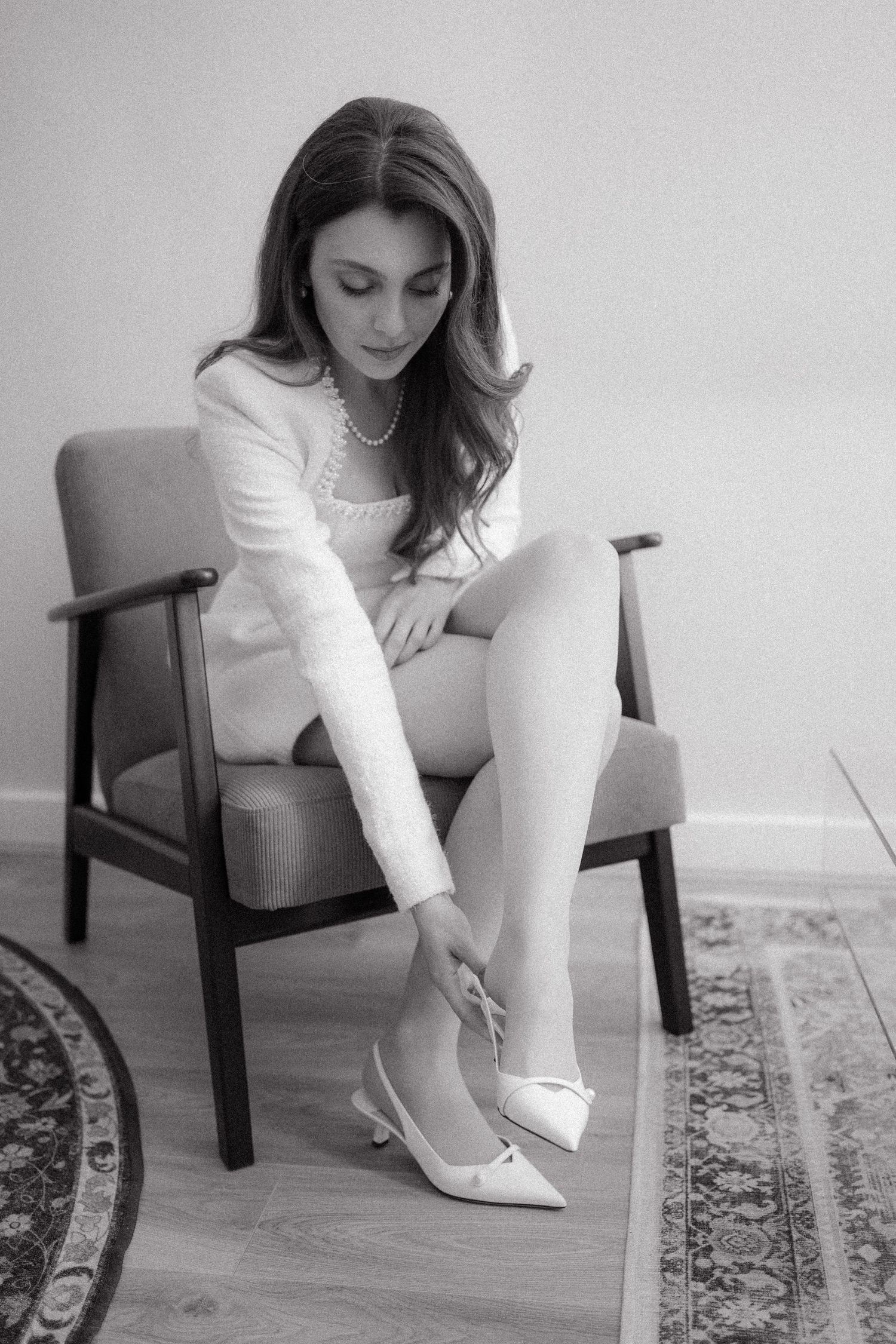 Black and white photograph showing white heels and dress details on a patterned rug.