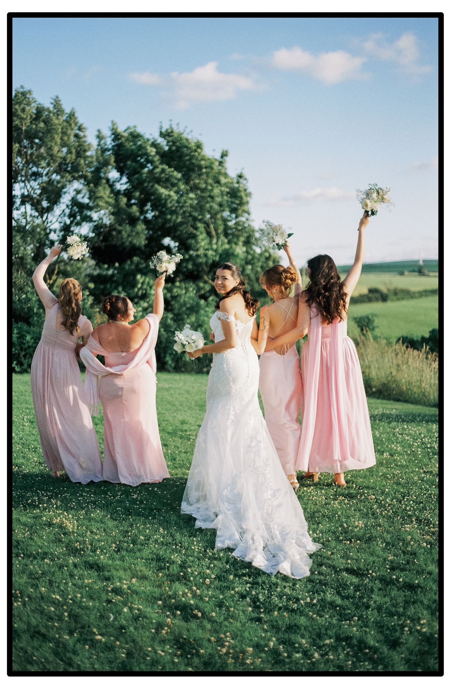 Wedding party in pink dresses dancing joyfully on a grassy hillside.