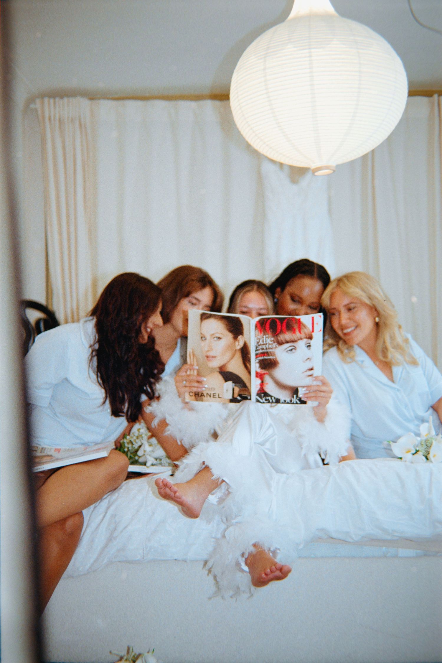 A group of friends in white robes relax together on a bed while reading magazines and laughing.