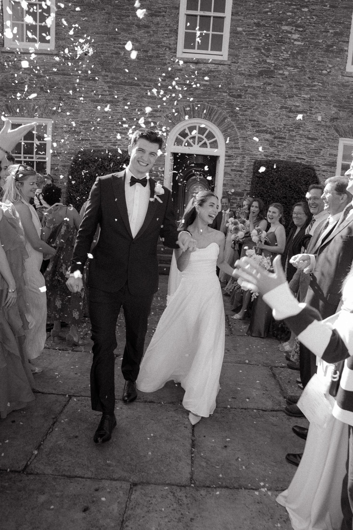 Wedding guests celebrate by throwing flower petals as newlyweds exit historic brick building during daytime ceremony.