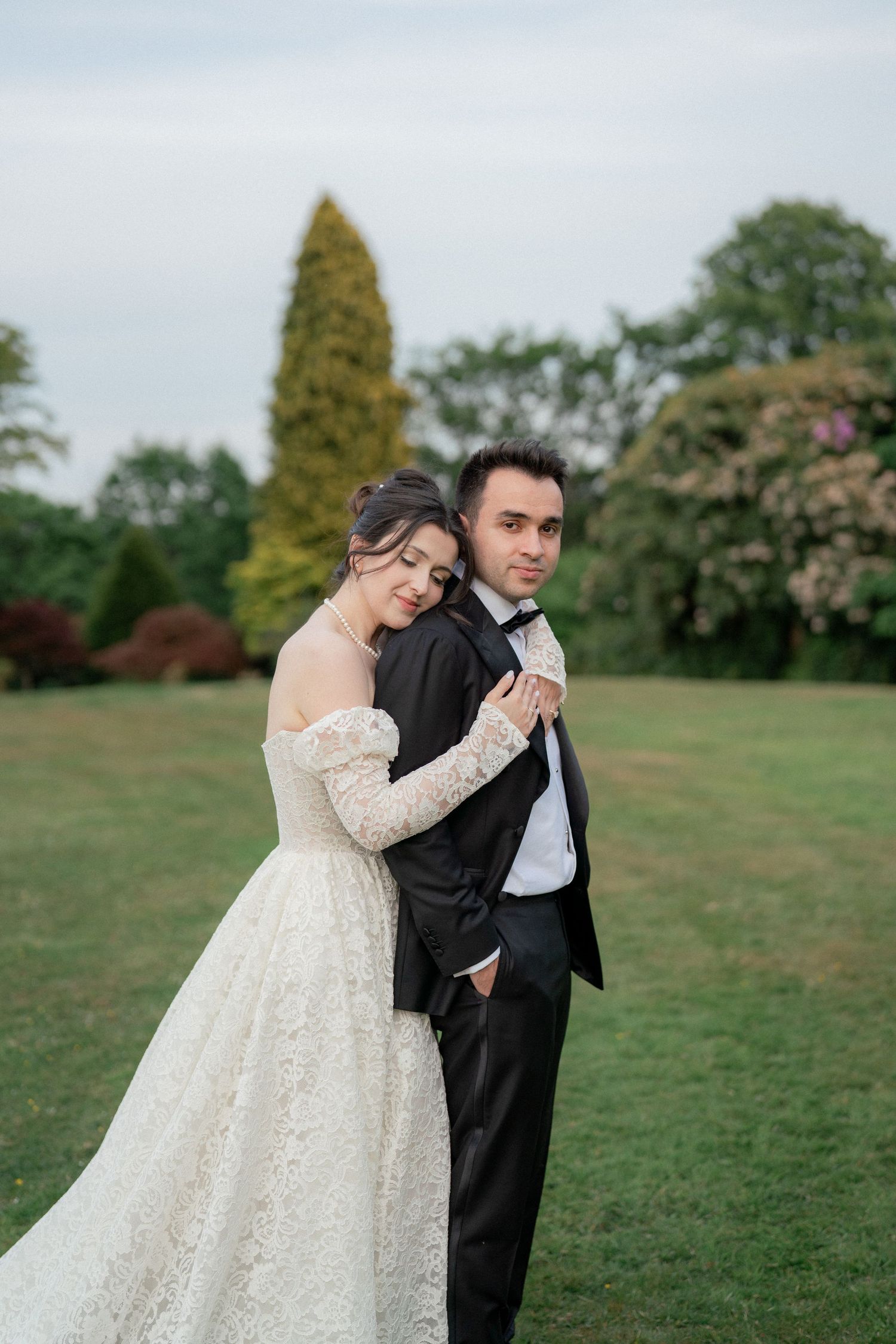 Romantic wedding couple embraces on a lush green lawn with trees and flowers in the background.