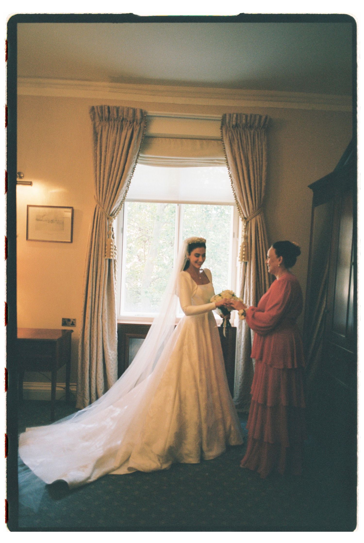 Bride in white gown with veil receives bouquet from woman in pink dress by curtained window.