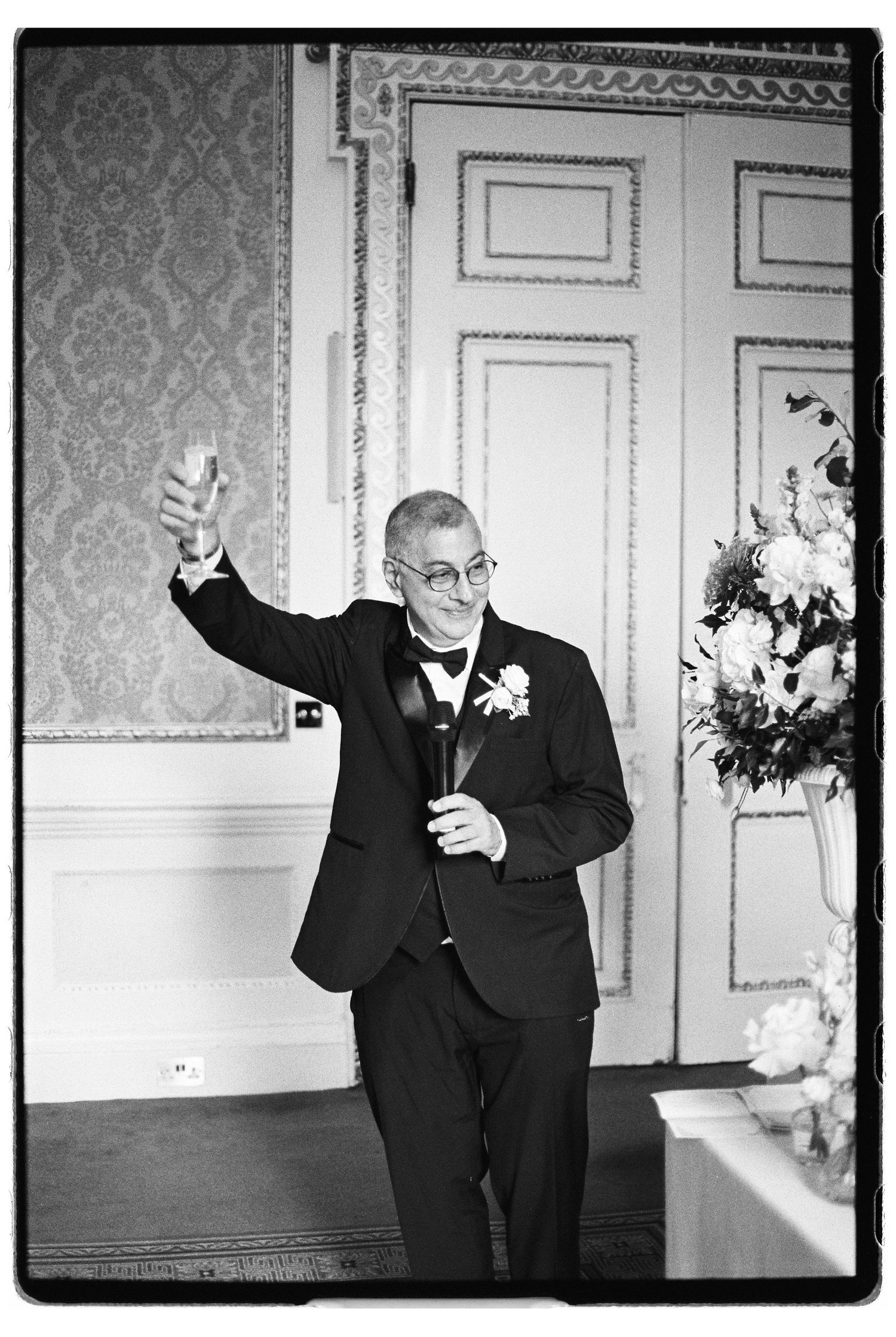 Man in tuxedo raises champagne glass giving a toast at wedding reception in black and white photo.