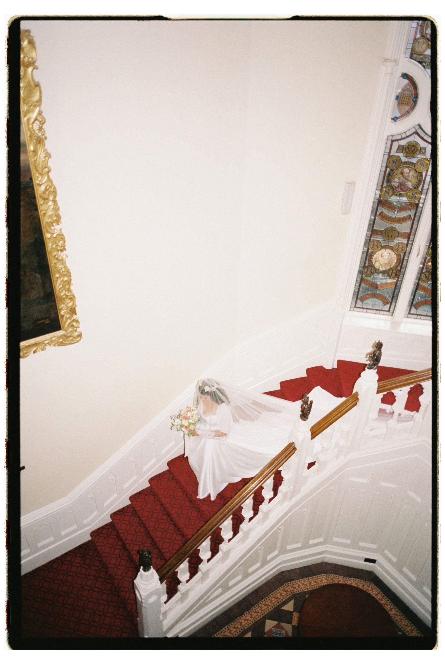 Bride in white gown descends grand staircase with red carpet holding bouquet.