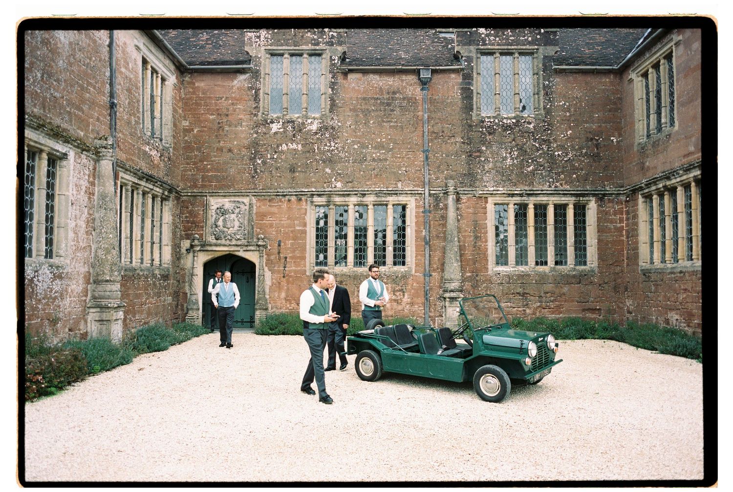 Groomsmen with a vintage green Mini Moke car outside a historic brick manor house wedding venue.
