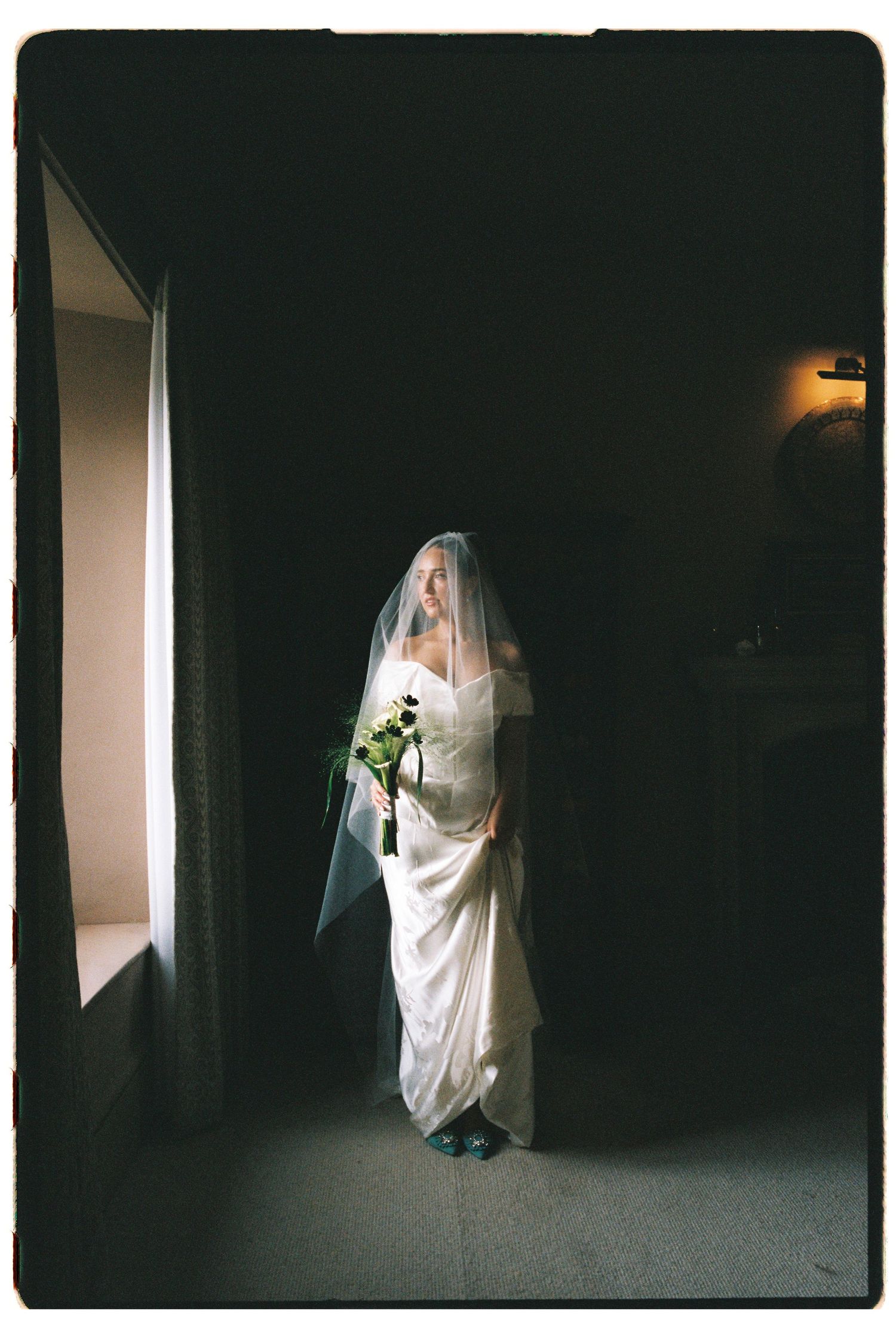 Bride in white gown and veil holding bouquet standing in dramatic light in a dark hallway.