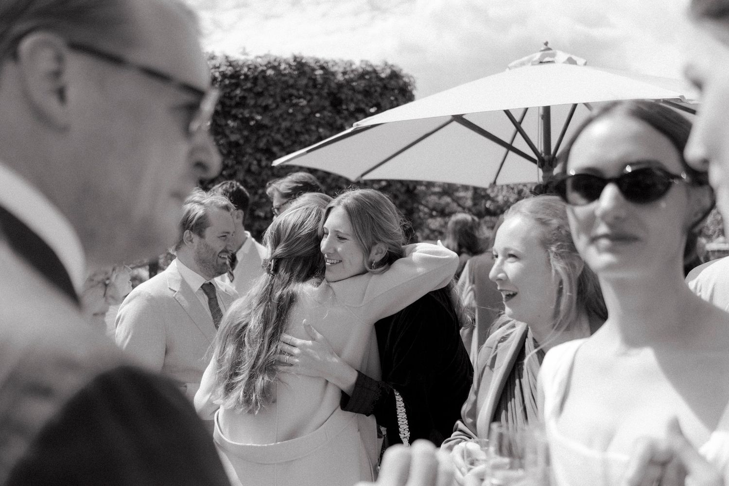 Wedding guests embrace and smile during an outdoor garden celebration beneath a white parasol, in a candid black and white photo.