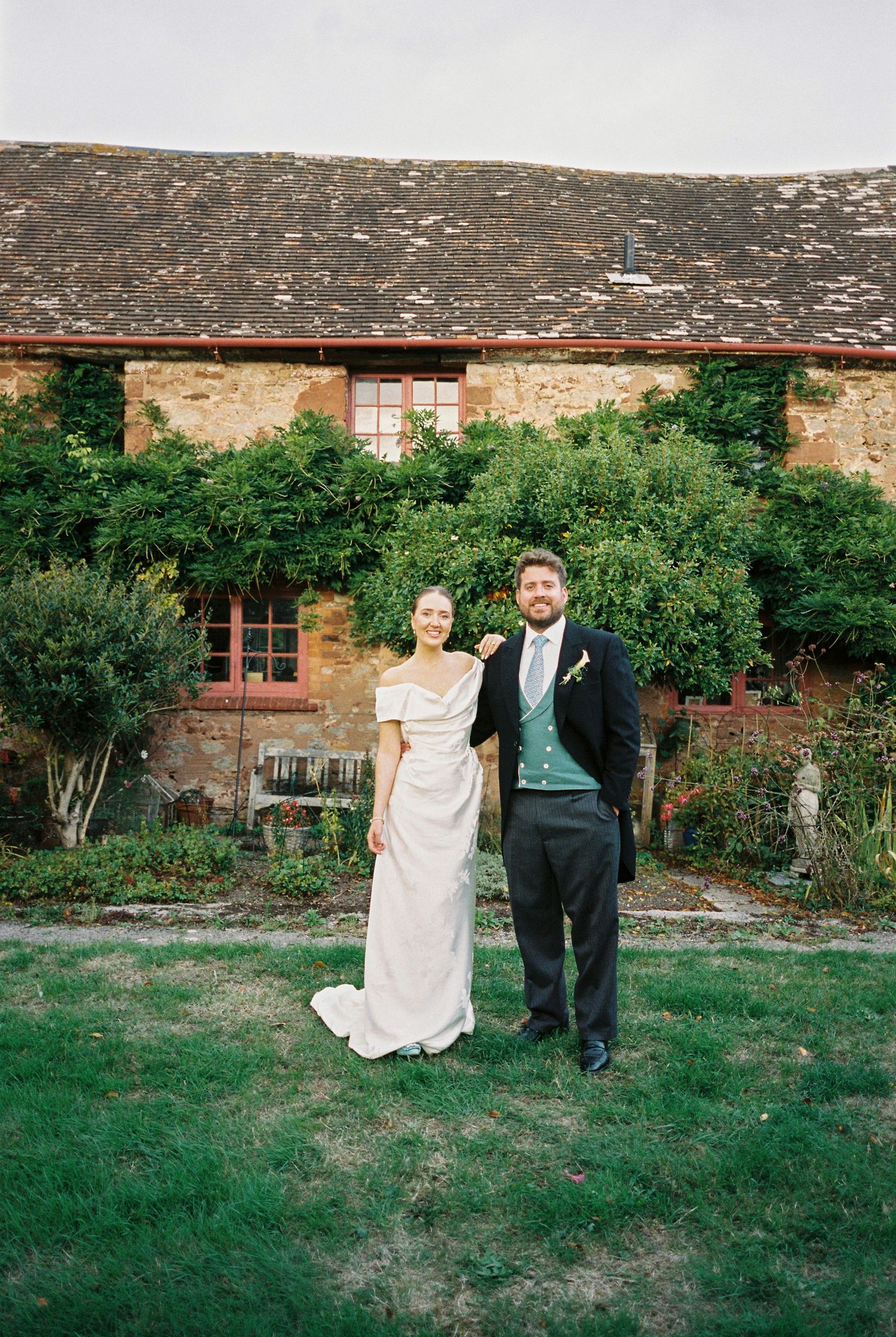 Bride in an off-shoulder ivory gown and groom in a dark suit with teal vest pose on a lawn before a vine-covered stone cottage.