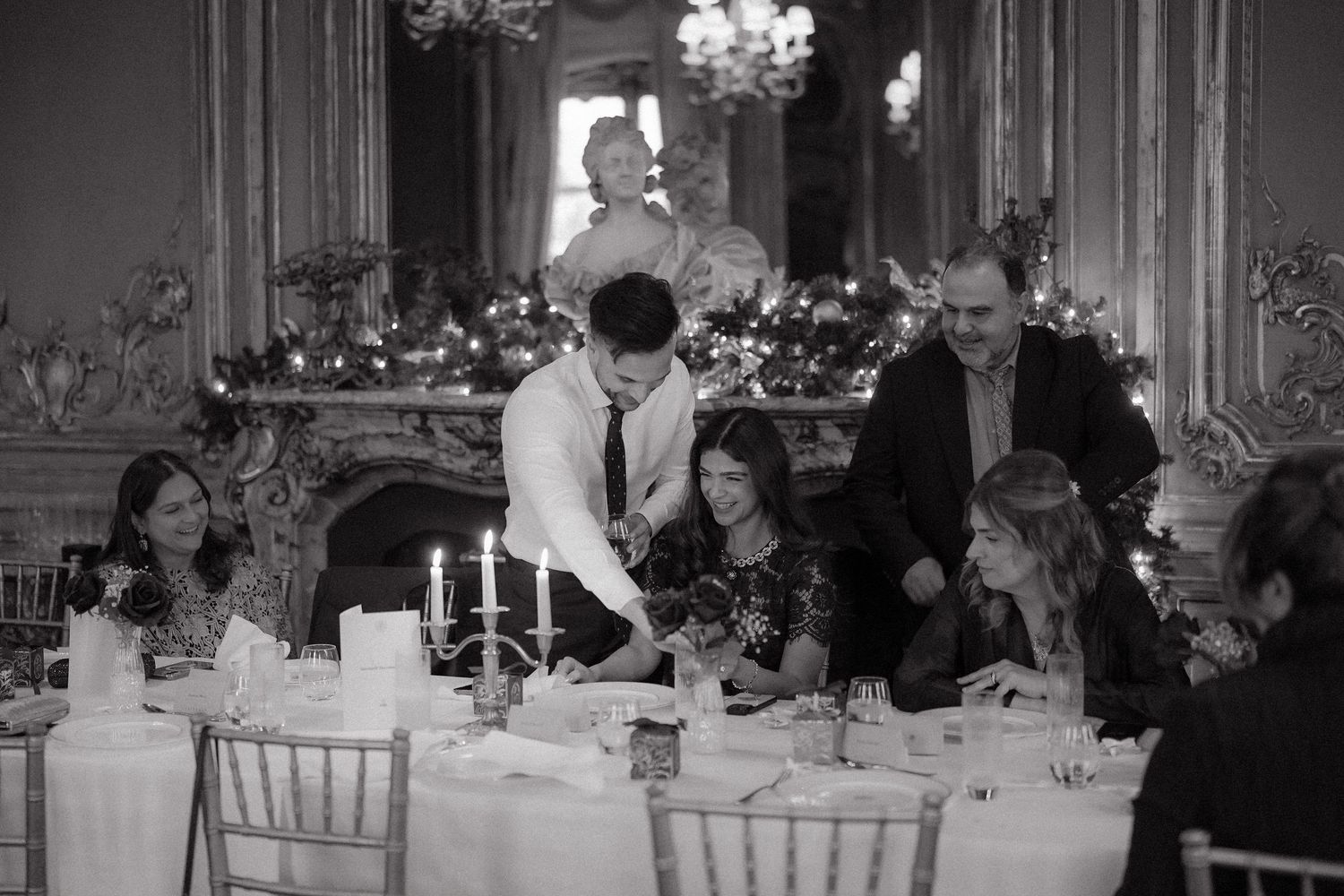 Elegant black and white photo of guests dining at a formal candlelit table in an ornate room with fireplace.