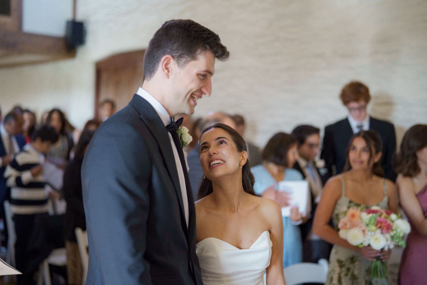 A joyful couple sharing a candid laughing moment during their wedding celebration with guests in the background.